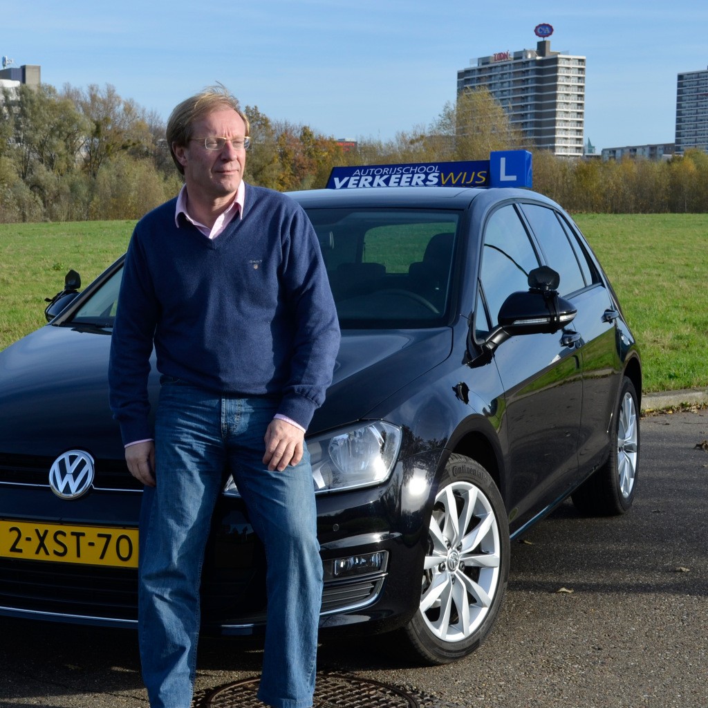 A smiling young man with crossed arms, wearing a plaid shirt and white t-shirt, poses against a dark background.