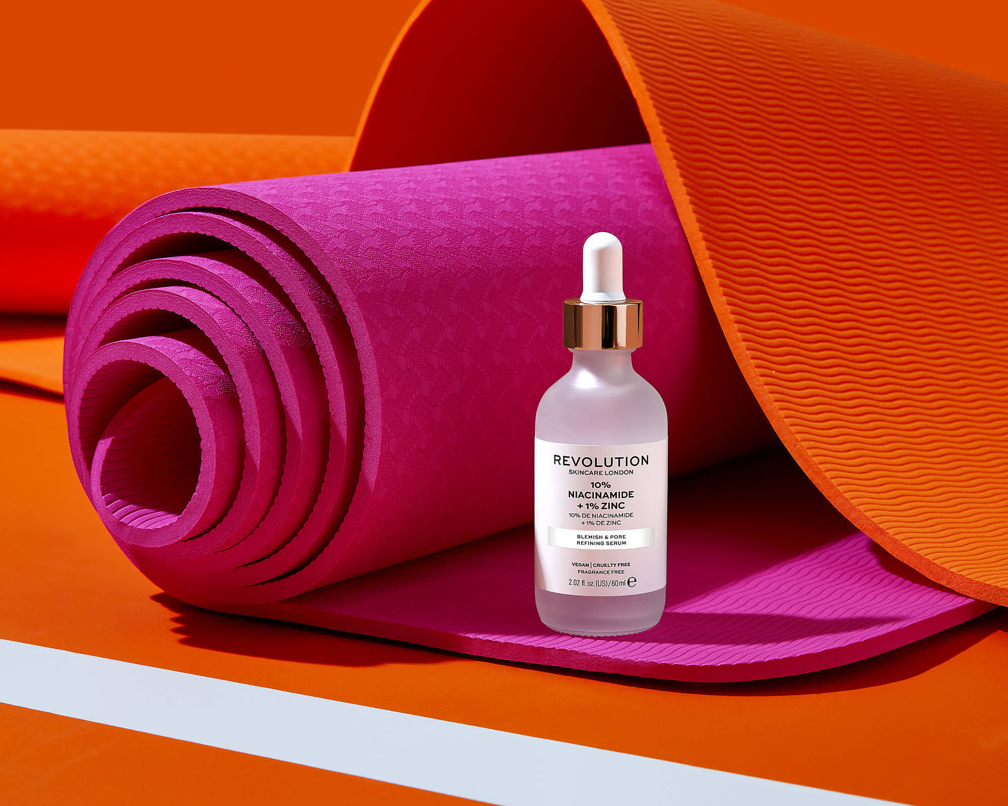 A clear bottle sits on a pink rolled fabric, with bright orange and white backgrounds creating a vibrant setting.
