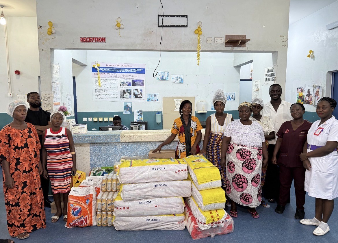 A man stands while two people sit at a table in an office setting, smiling and engaged in conversation. The mood is friendly and collaborative.