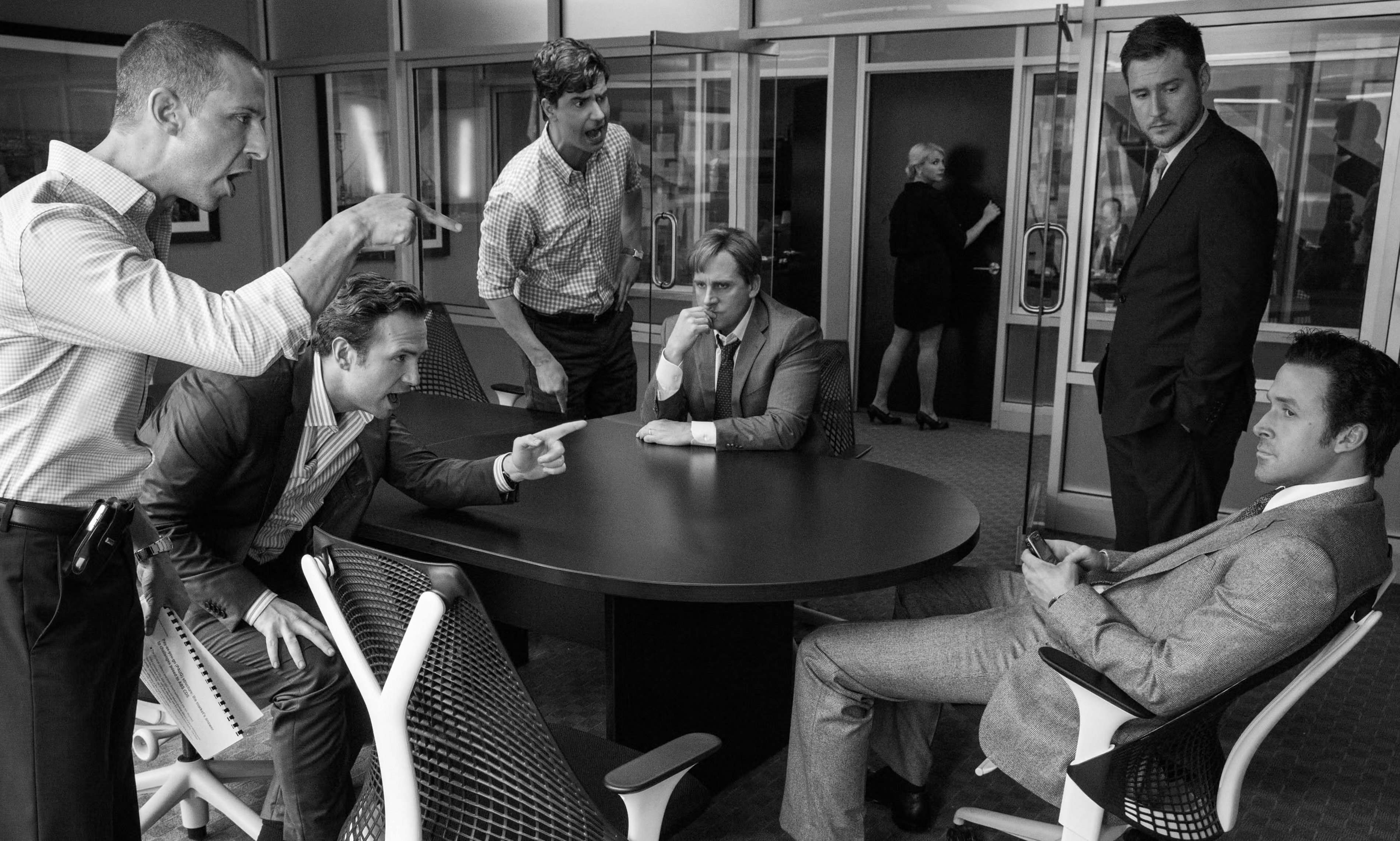 Several men in business attire are engaged in a heated argument, with some standing and aggressively pointing fingers, while others sit, observing or reacting. One man in a grey suit, seated and leaning back, appears calm, holding a phone. The setting features a modern glass-walled conference room with a dark wooden table and ergonomic office chairs. The scene conveys tension, conflict, and high-stakes financial discussions.