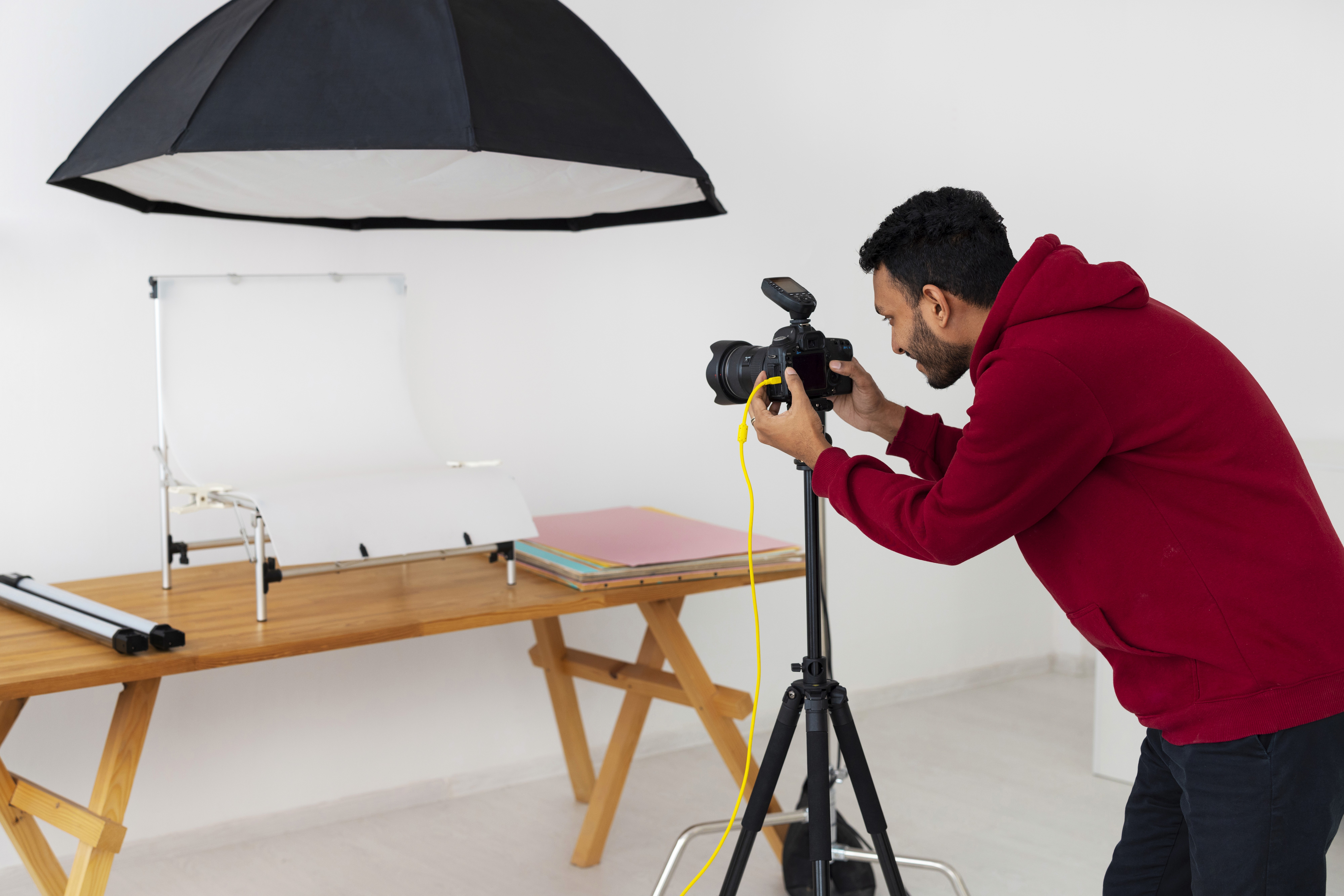 Man in a red hoodie photographing products on a wooden table using a DSLR camera mounted on a tripod in a studio setup with softbox lighting and white backdrop.