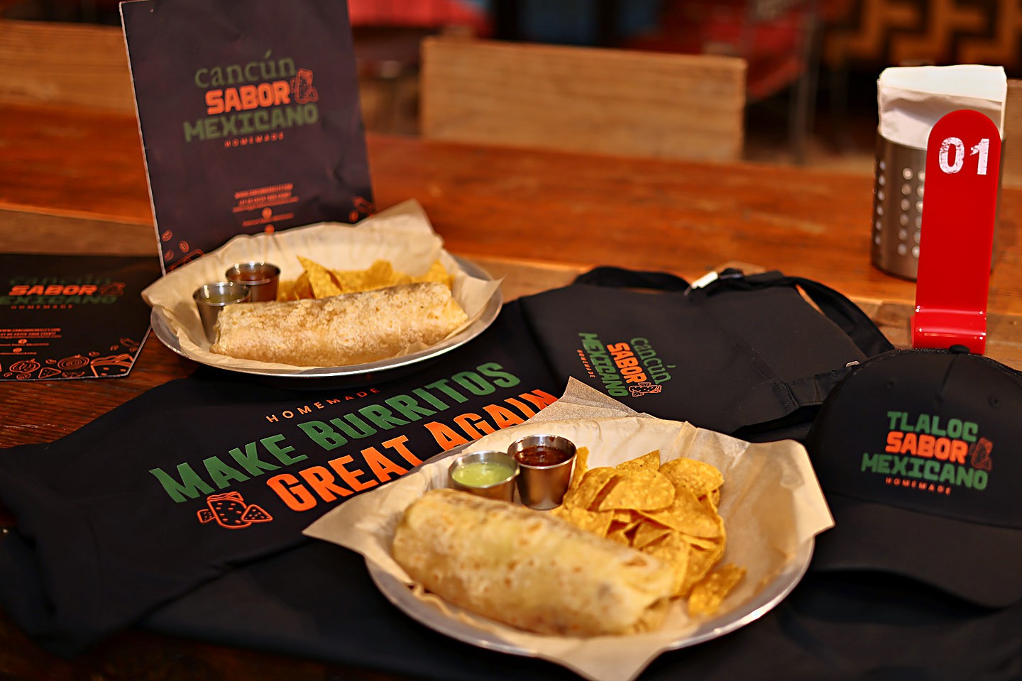 Two burrito plates on a wooden table with the branded table tent, the printed tee, a tote bag, and the cap