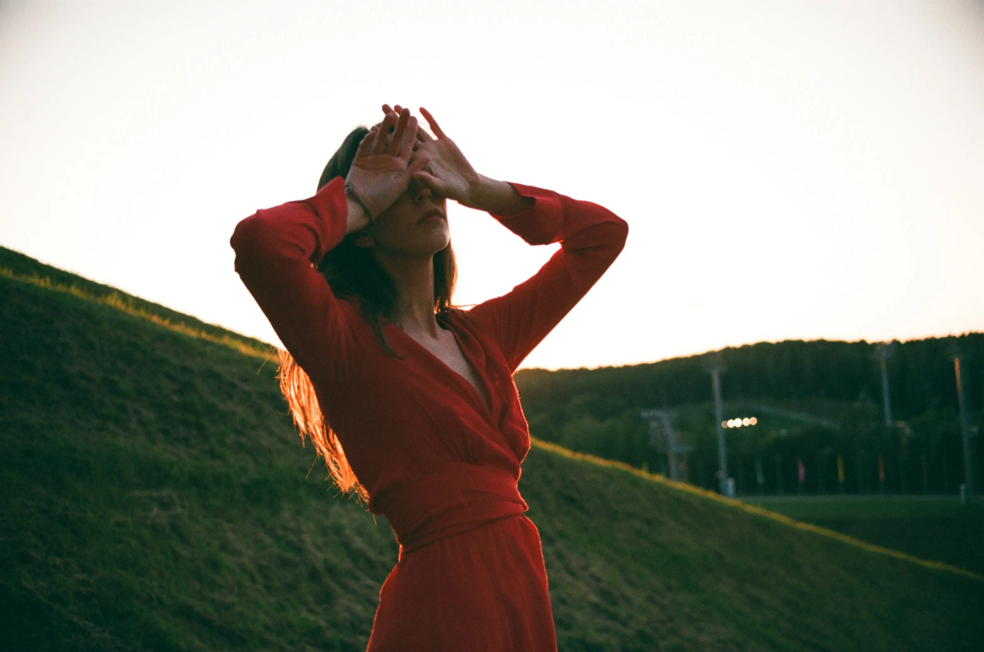 Woman in red dress standing in the field, hands resting on her eyes.