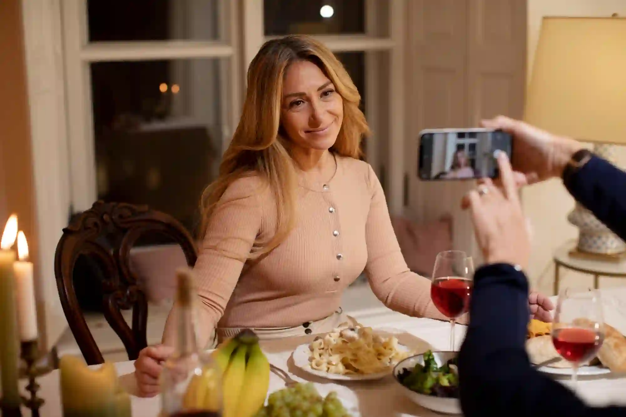 A smiling woman at a dinner table is being photographed by someone holding a smartphone.