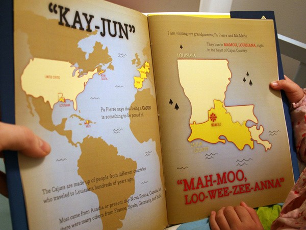 Two hands hold open an open children’s book titled “KAY-JUN” on the left page and “MAH-MOO, LOO-WEE-ZEE-ANNA” on the right page. The left page shows a map of the world with text describing the Cajuns and their migration from various countries. The right page displays a map of Louisiana with text mentioning the Grandparents’ visit to MAMOU, Louisiana. The book’s pages are cream-colored with dark blue edges.