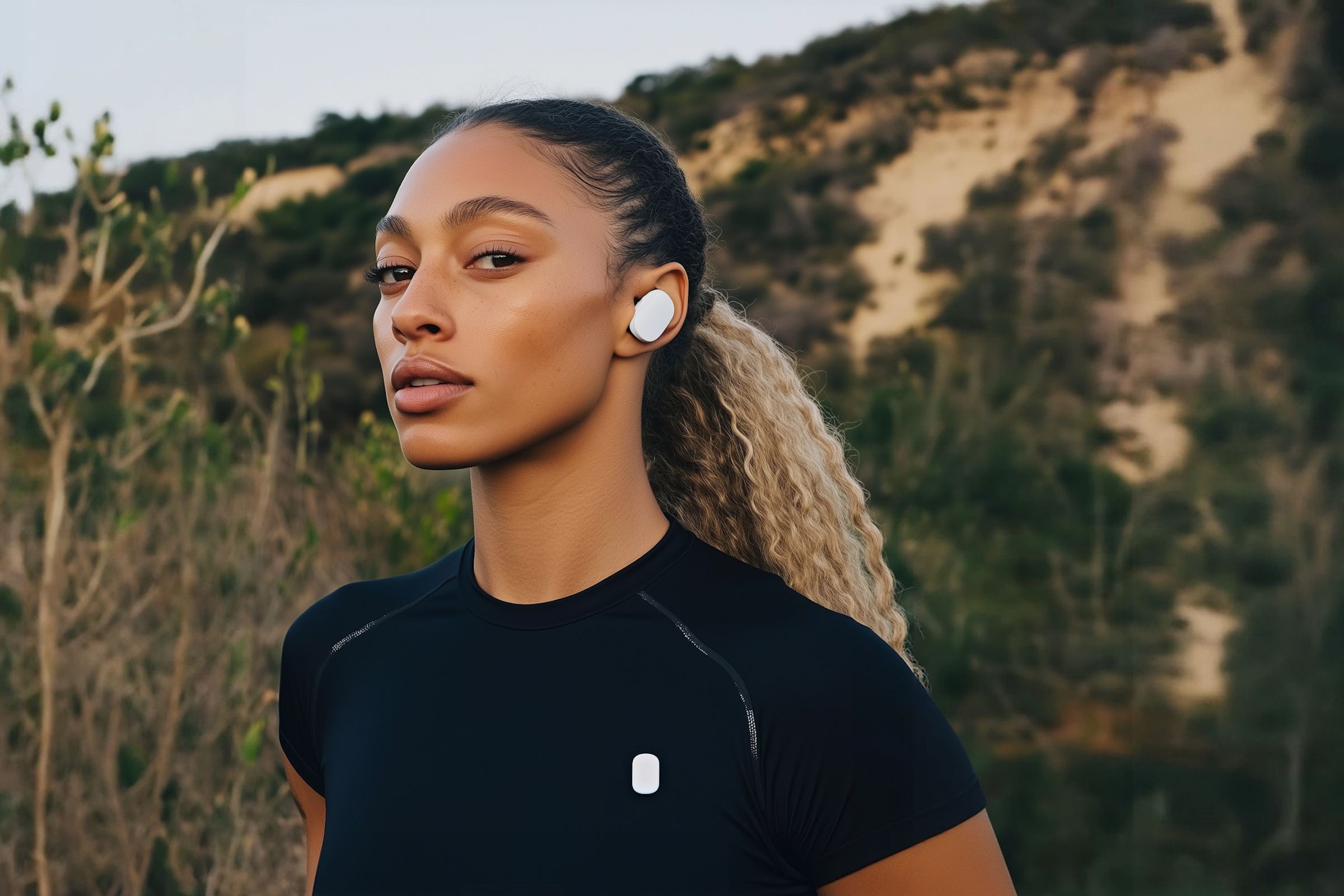 A woman in athletic wear wearing white wireless earbuds outdoors, with a blurred natural landscape in the background.