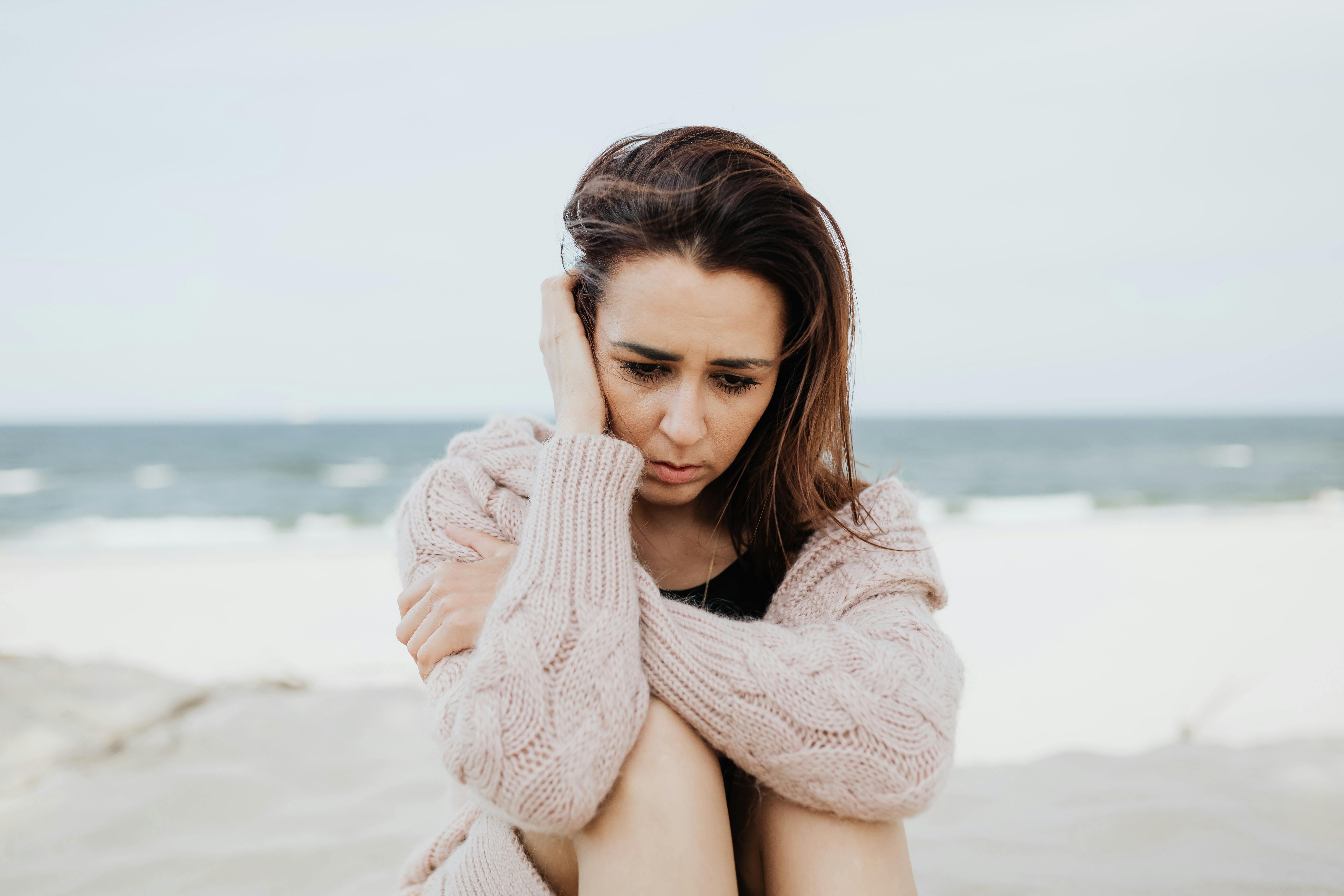 A woman looking scared and worried is seated on the beach holding her head in her arms