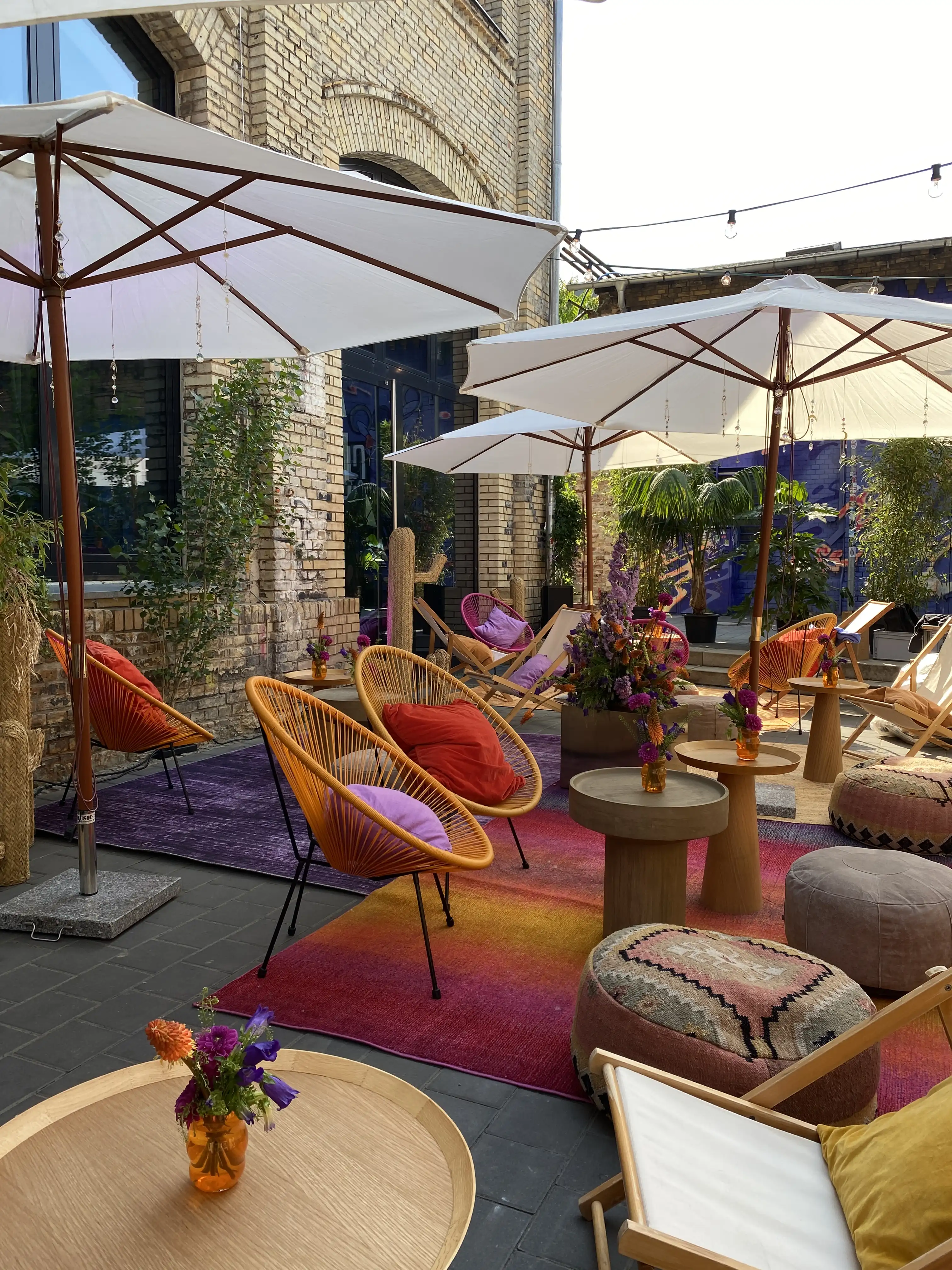 Sunny courtyard lounge with orange acapulco chairs, parasols, poufs and colourful gradient rugs.