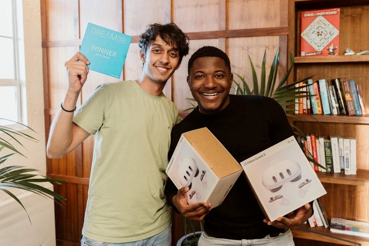 Two smiling men standing together indoors, one holding a blue ‘DreamXR Winner’ sign and the other holding two boxed Meta Quest 3 headsets. They appear happy and excited, celebrating their achievement. The background features wooden panels, a bookshelf with various books, and a green plant.