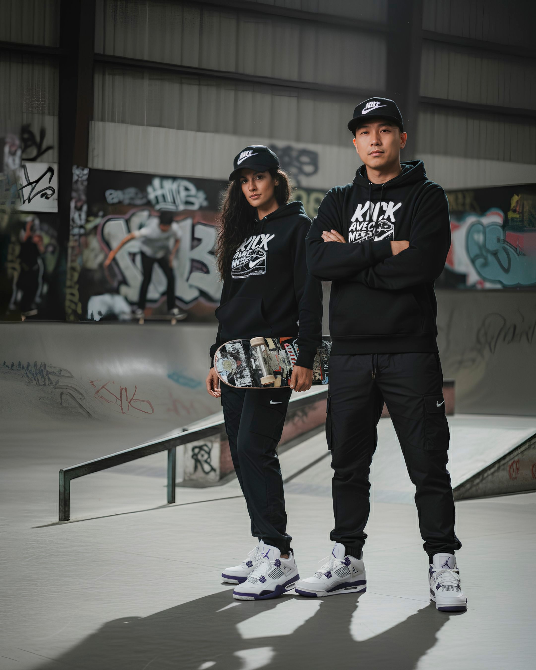 Photo de mode lifestyle d'un duo (homme et femme) posant en tenue streetwear coordonnée dans un skatepark couvert et graffité. Les deux modèles portent des casquettes noires 'Nike', des sweats à capuche noirs avec un imprimé graphique blanc, et des pantalons de survêtement/cargo noirs, ainsi que des sneakers blanches et violettes (Jordan 4). La femme tient une planche de skateboard. L'homme est photographié les bras croisés, adoptant une posture confiante. L'arrière-plan flou, avec des skateurs en action, souligne la thématique culture urbaine.