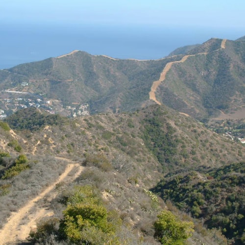 一条蜿蜒的土路穿过丘陵的干旱景观，稀疏的植被点缀其间，引向远方的群山和朦胧的地平线。