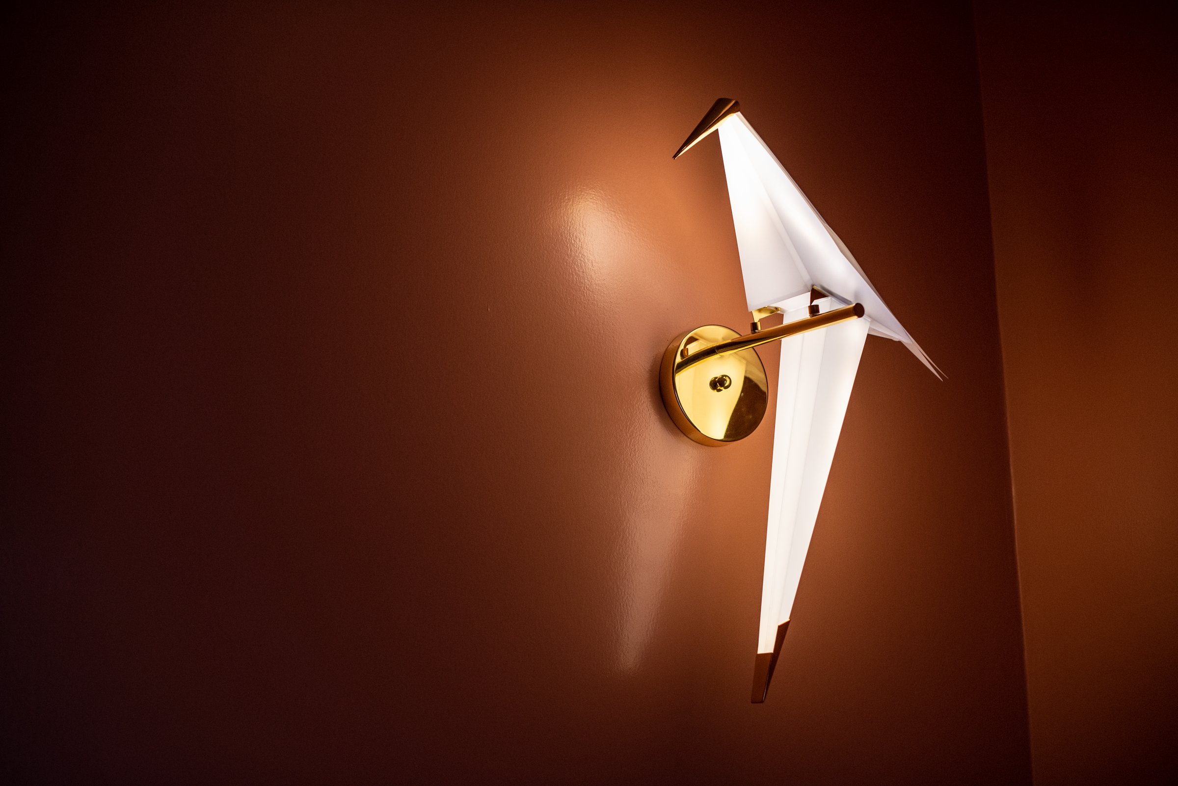 Angular white bird-shaped wall light with brass fittings against a deep reddish-brown wall.