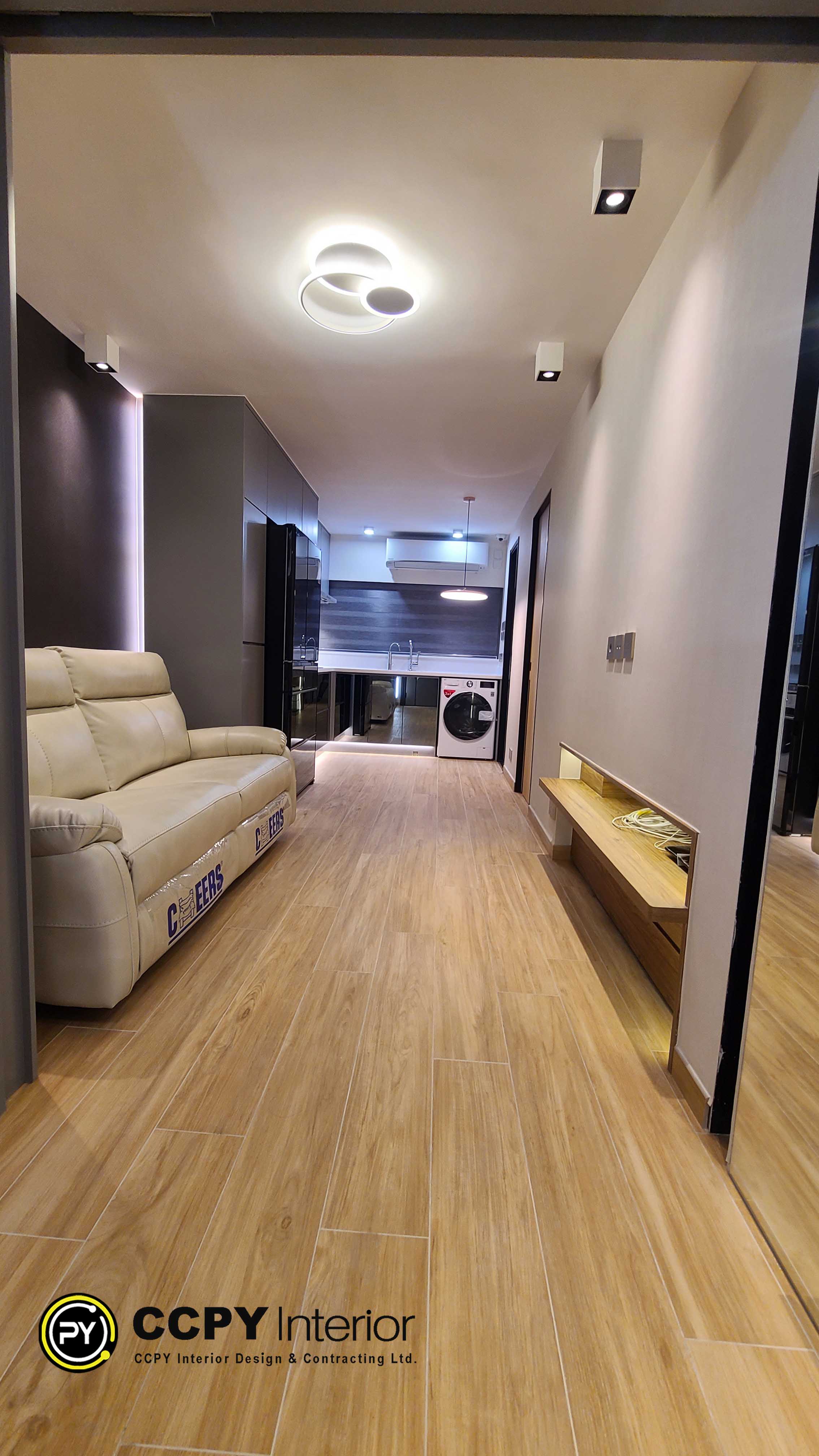 The selected region shows an interior view of a long, narrow residential apartment designed with modern, space-saving solutions suitable for Hong Kong homes.Key Features of the SpaceLayout: A long, narrow galley configuration connecting the living area directly to the kitchen and utility space.Living Area: Features a large, light beige leather sofa and a wall-mounted wooden console unit on the opposite side to maximize floor space.Flooring: Light-colored wood-look laminate or vinyl planks running lengthwise to make the narrow corridor appear longer.Kitchen & Utility: Located at the far end, featuring built-in cabinetry, a dark mirrored refrigerator panel, and an integrated front-loading washing machine under the counter.