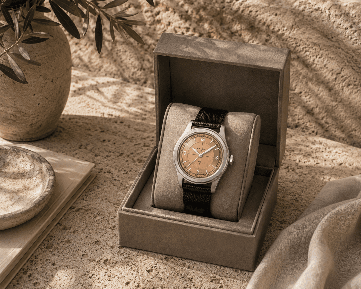 A Baltic MR01 stainless steel watch with a salmon-colored sector dial and black leather strap showcased inside an open taupe presentation box. The watch box sits on a textured travertine stone ledge next to a ceramic vase with olive foliage, a small stone dish resting on a beige notebook, and draped linen fabric under warm, dappled sunlight.