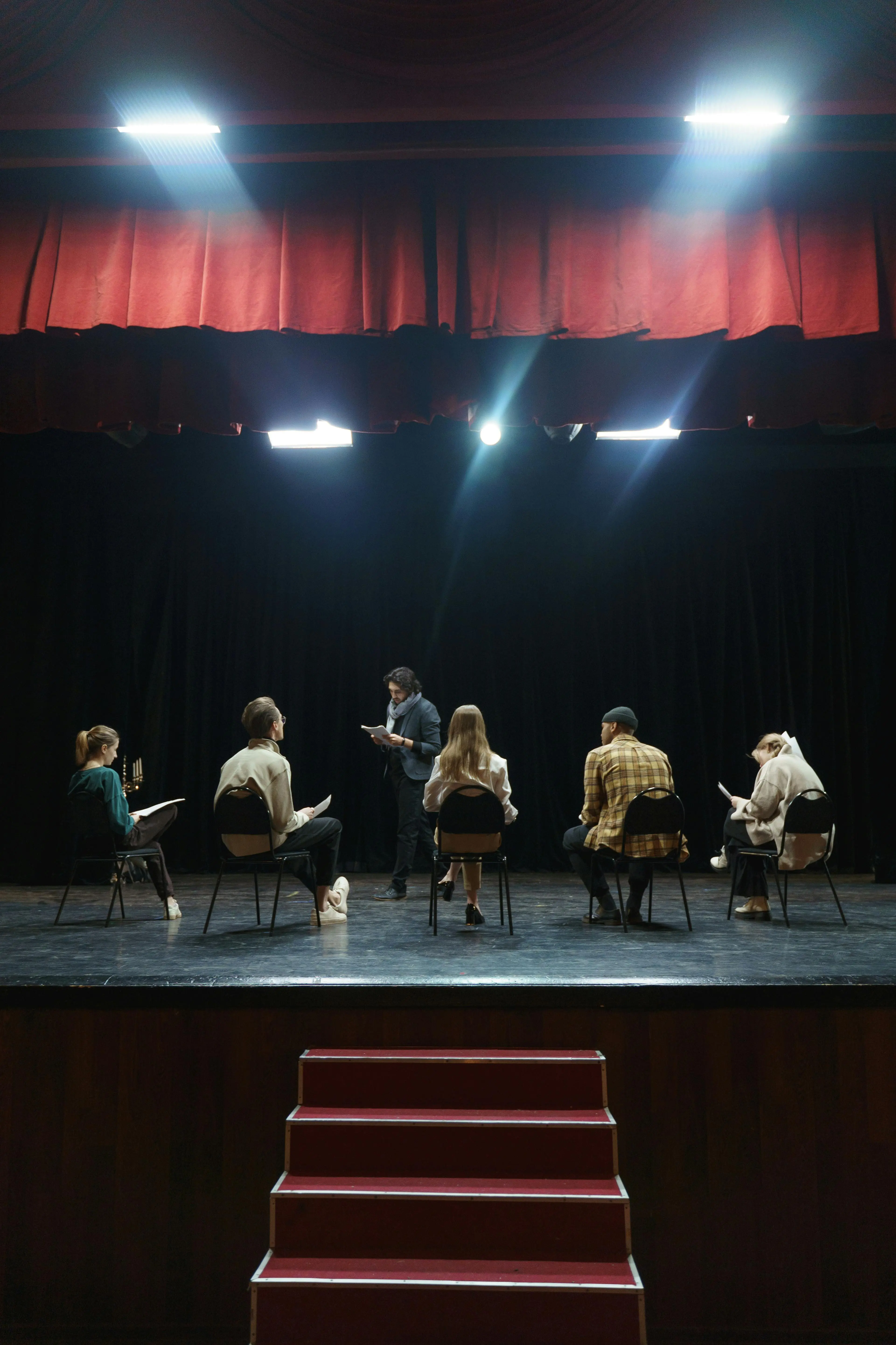 Theatre director standing on stage facing a semi-circle of seated young actors during auditions, symbolizing the collaborative and creative spirit of student theatre festivals.