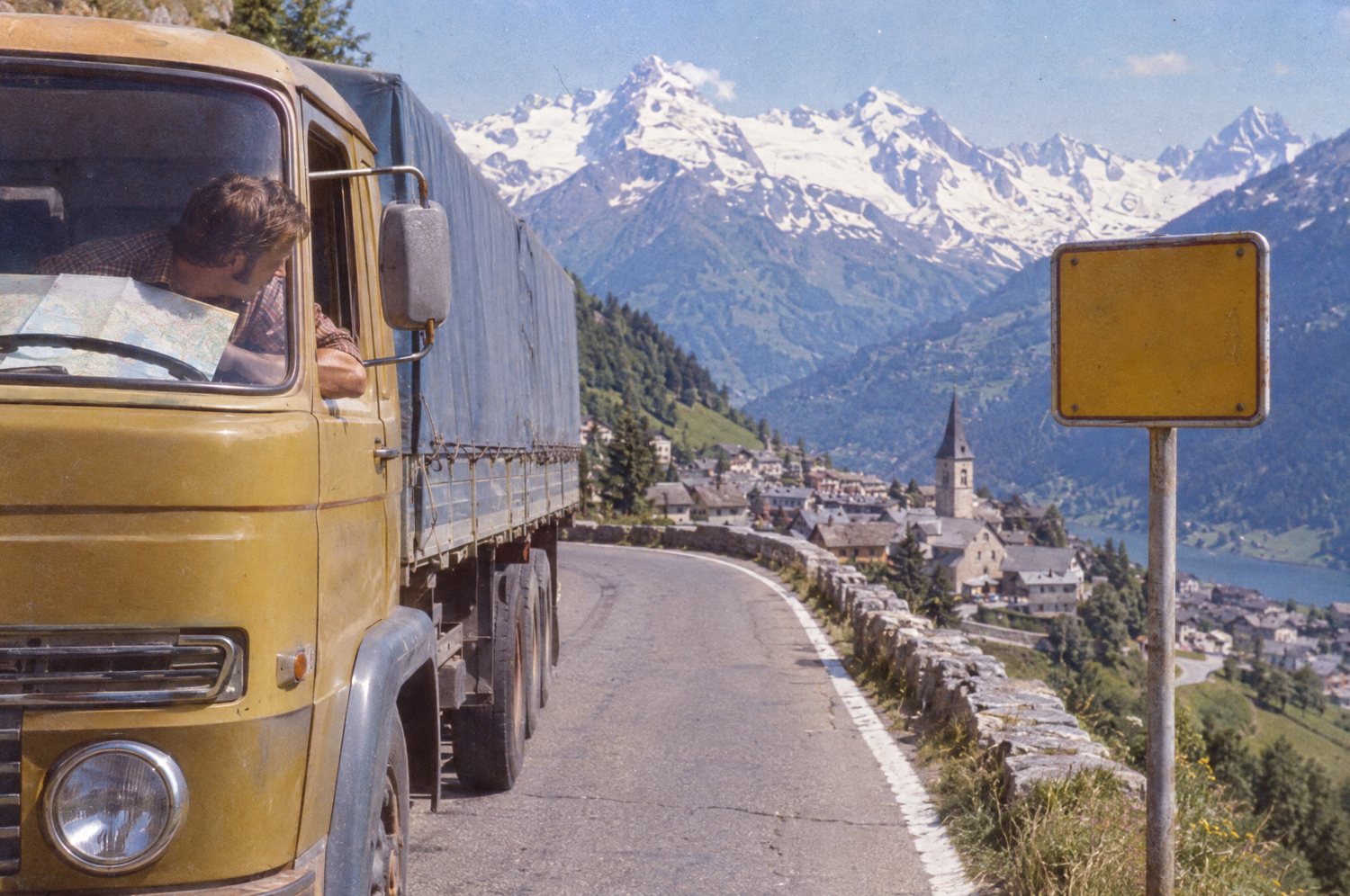 A lorry driver with a map leans out of his cab beside a blank yellow road sign above the town