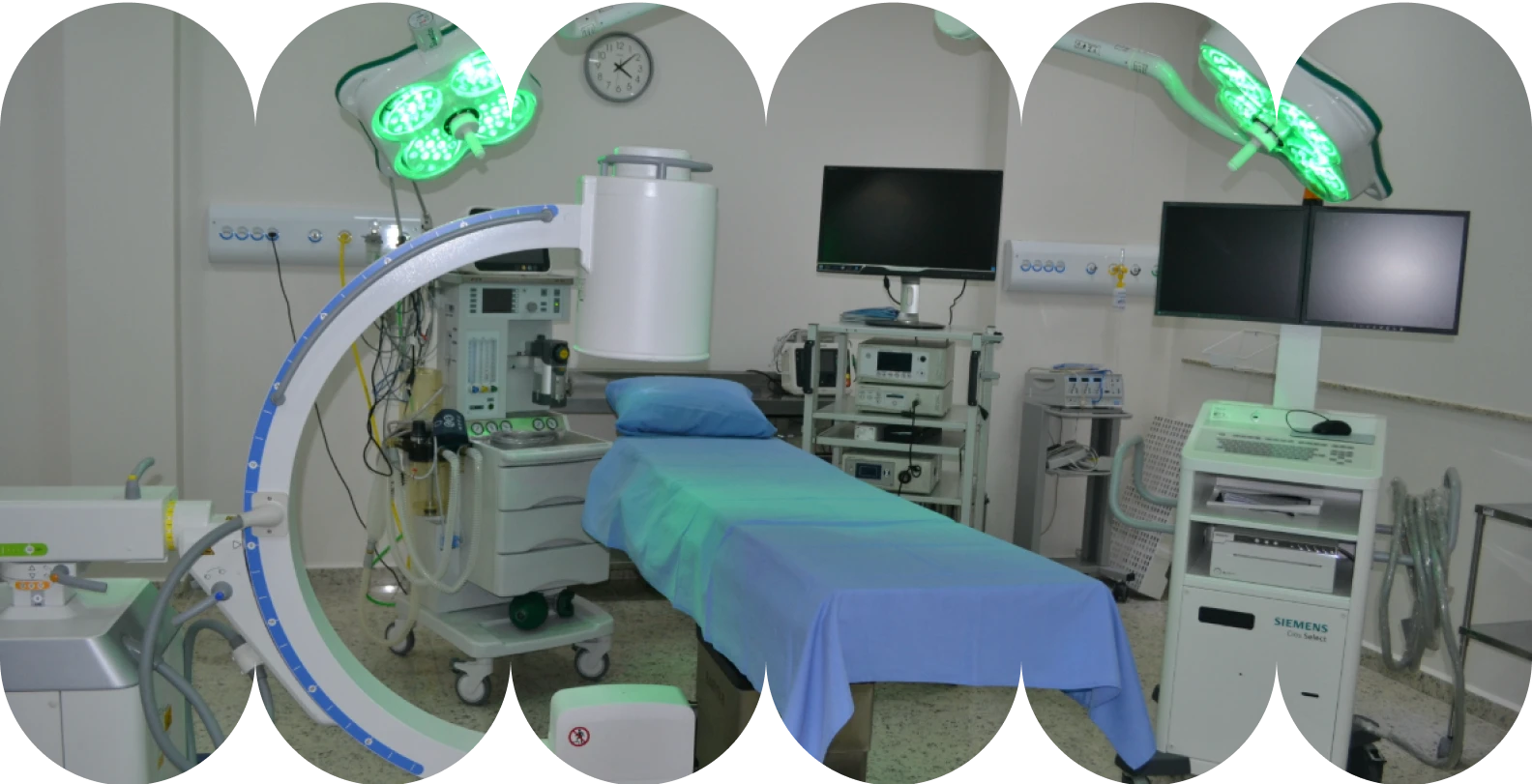 Imagem de uma sala de cirurgia moderna e bem equipada em um Hospital-Dia, mostrando foco na mesa de cirurgia coberta de azul, foco no aparelho de fluoroscopia em forma de C (arco cirúrgico) e em monitores e luminárias cirúrgicas.