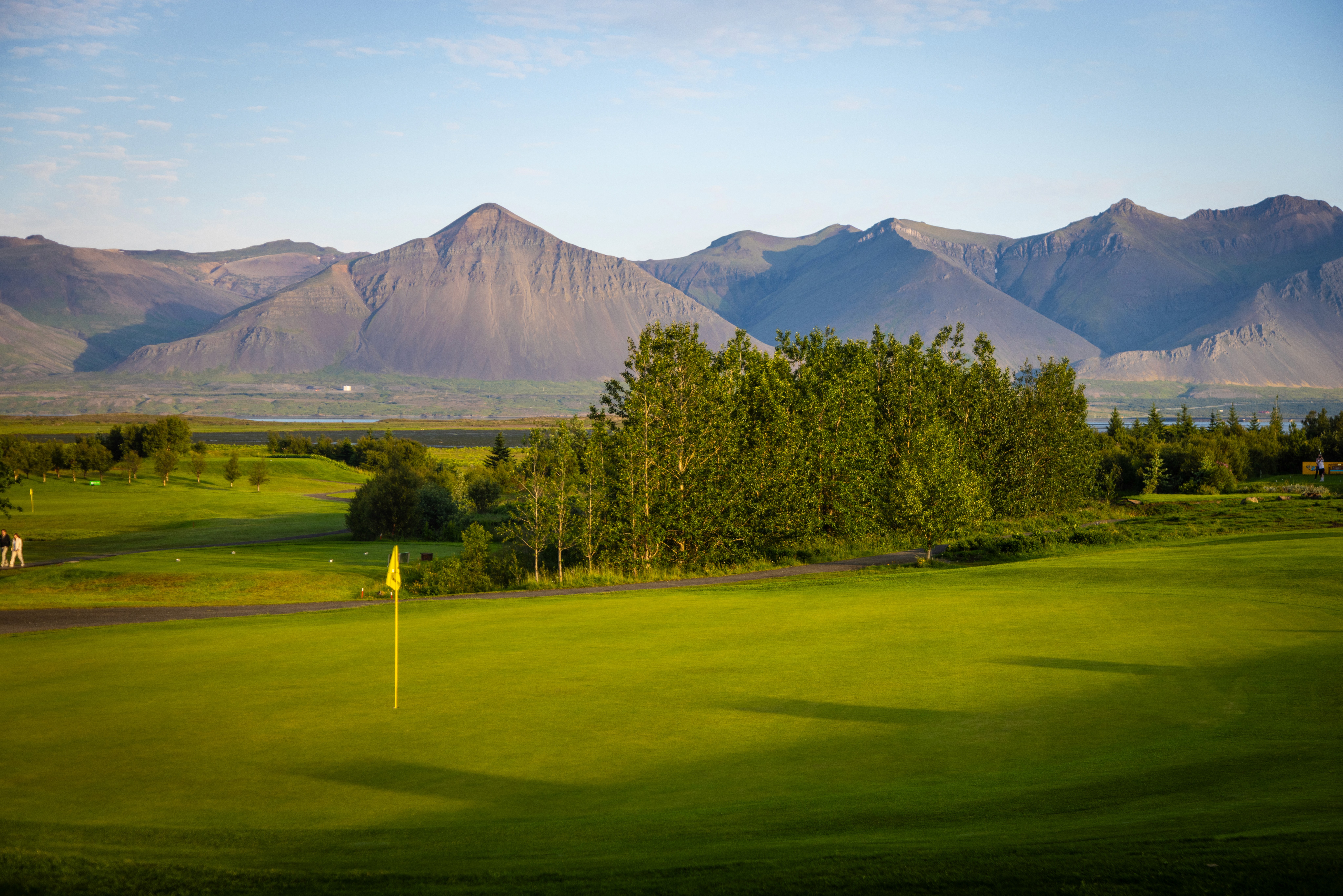 Gróskumikill grænn golfvöllur með gulum fána í forgrunni, röð af háum trjám og stór, grýtt fjöll í bakgrunni undir blárri himni.