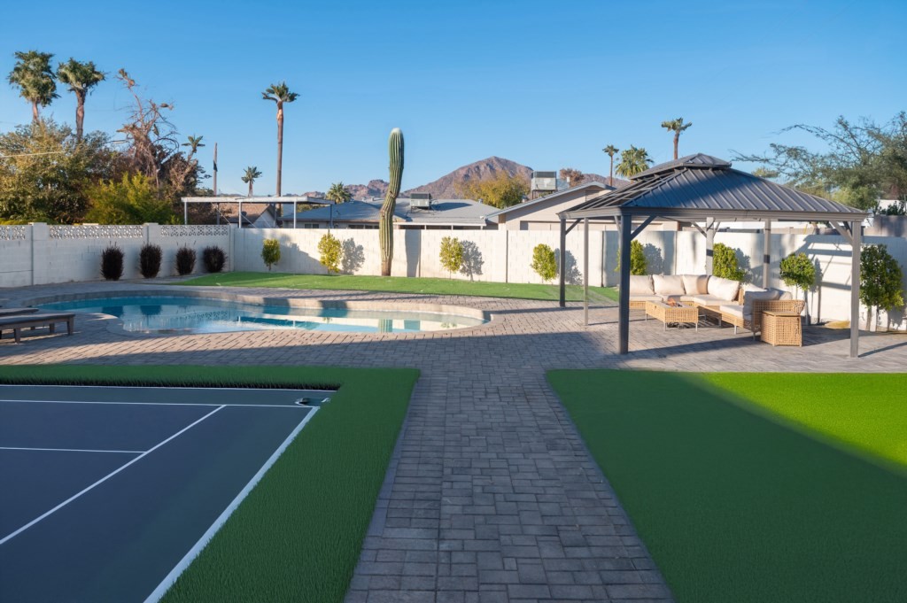 A spacious backyard features a modern pool, tennis court, and a shaded seating area under a gazebo, set against a backdrop of palm trees and a mountain view, epitomizing an affordable luxury vacation rental house.