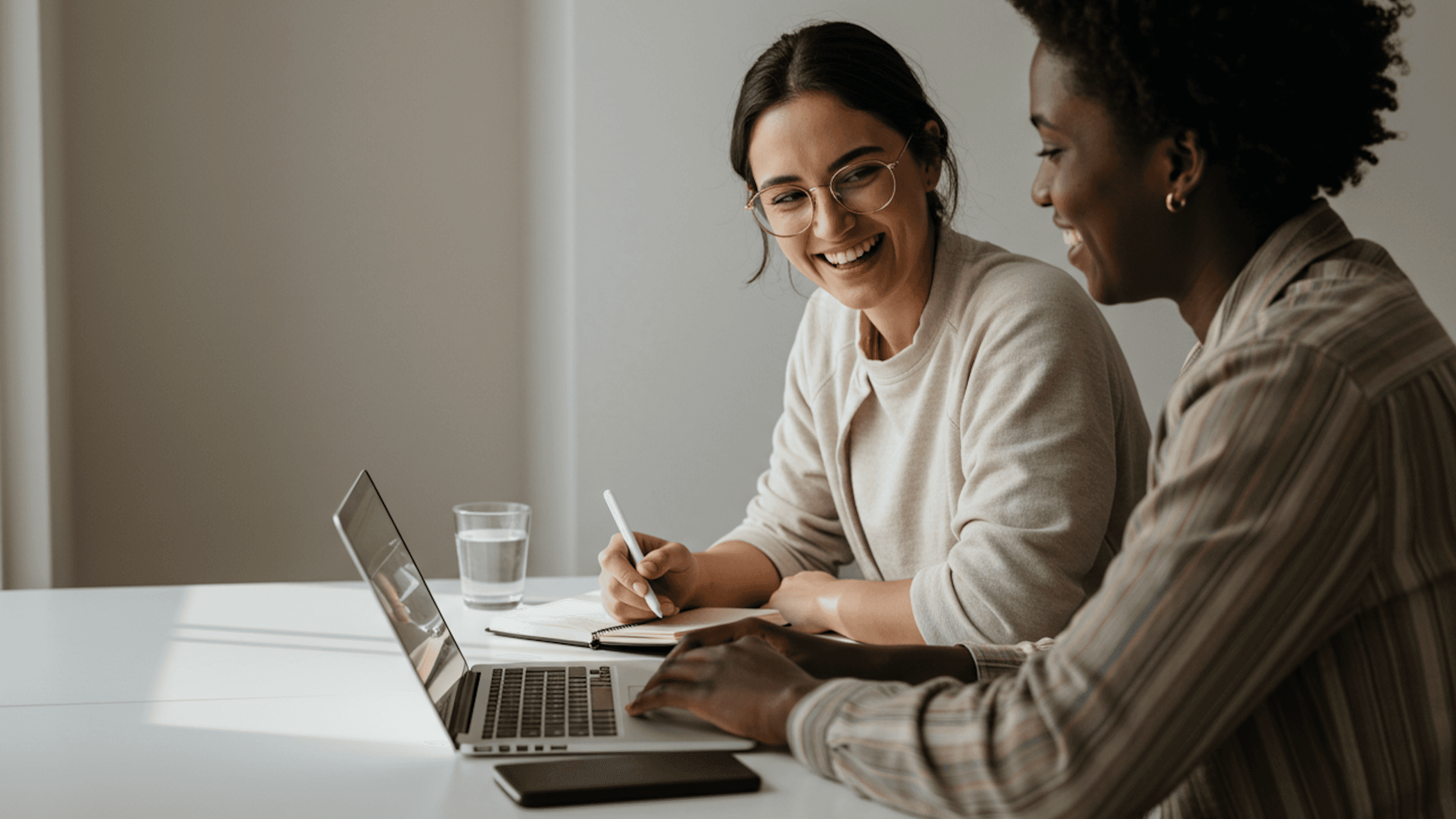 Two women collaborating at a table with a laptop, notebook, and glass of water, sharing ideas in a bright, modern workspace.