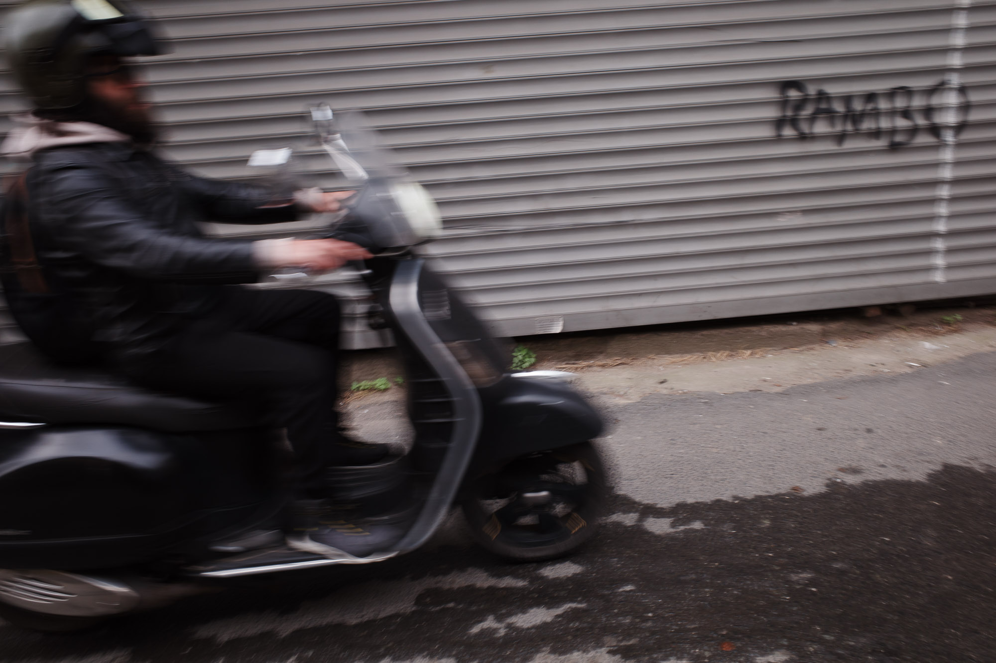 A person wearing a helmet speeds past on a black scooter, with a blurred motion effect, in front of a corrugated metal wall featuring graffiti.