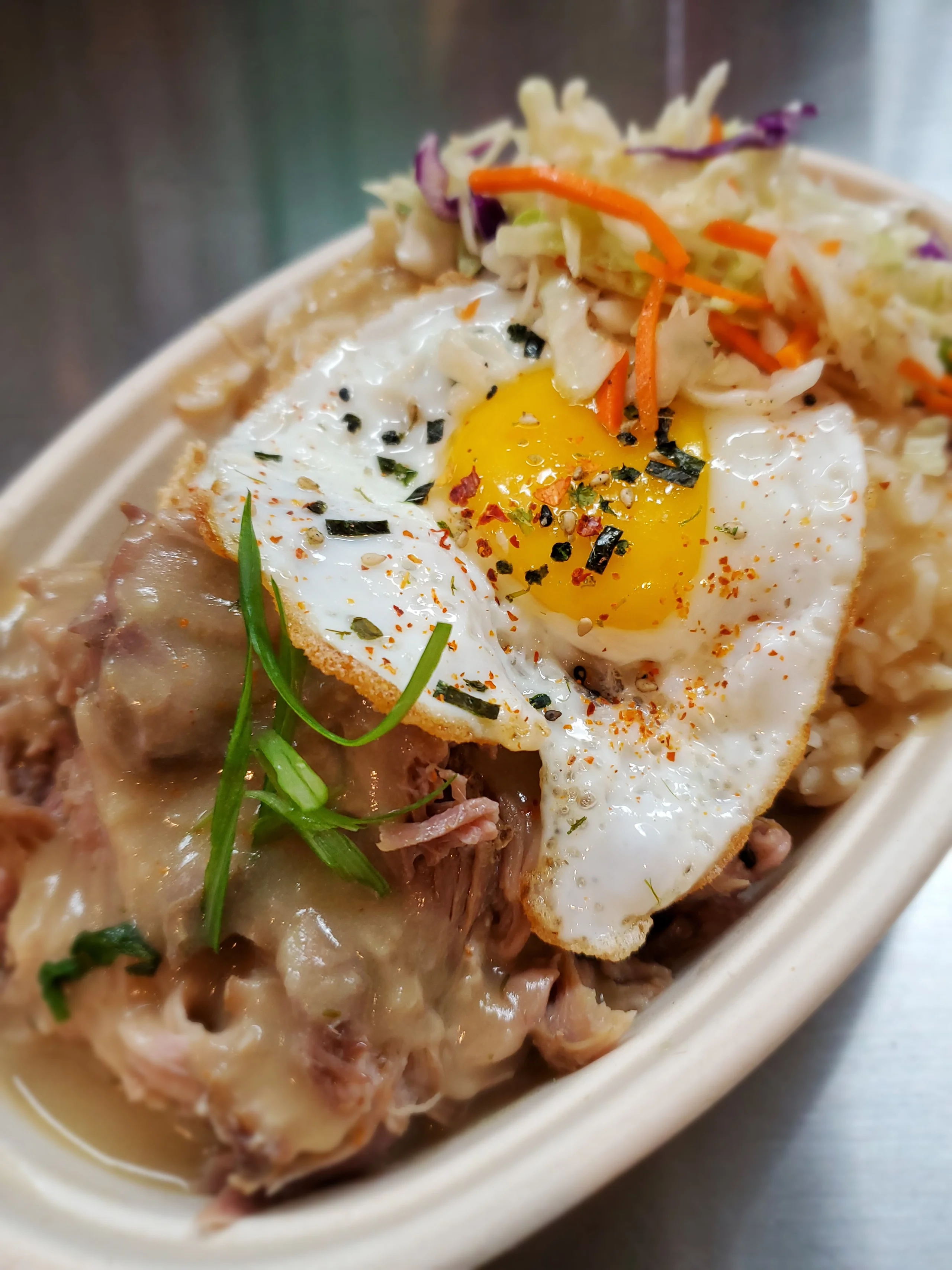 A bed of white rice topped with shredded Kalua pork, rich brown gravy, a fried egg, and black and green furikake seasoning.
