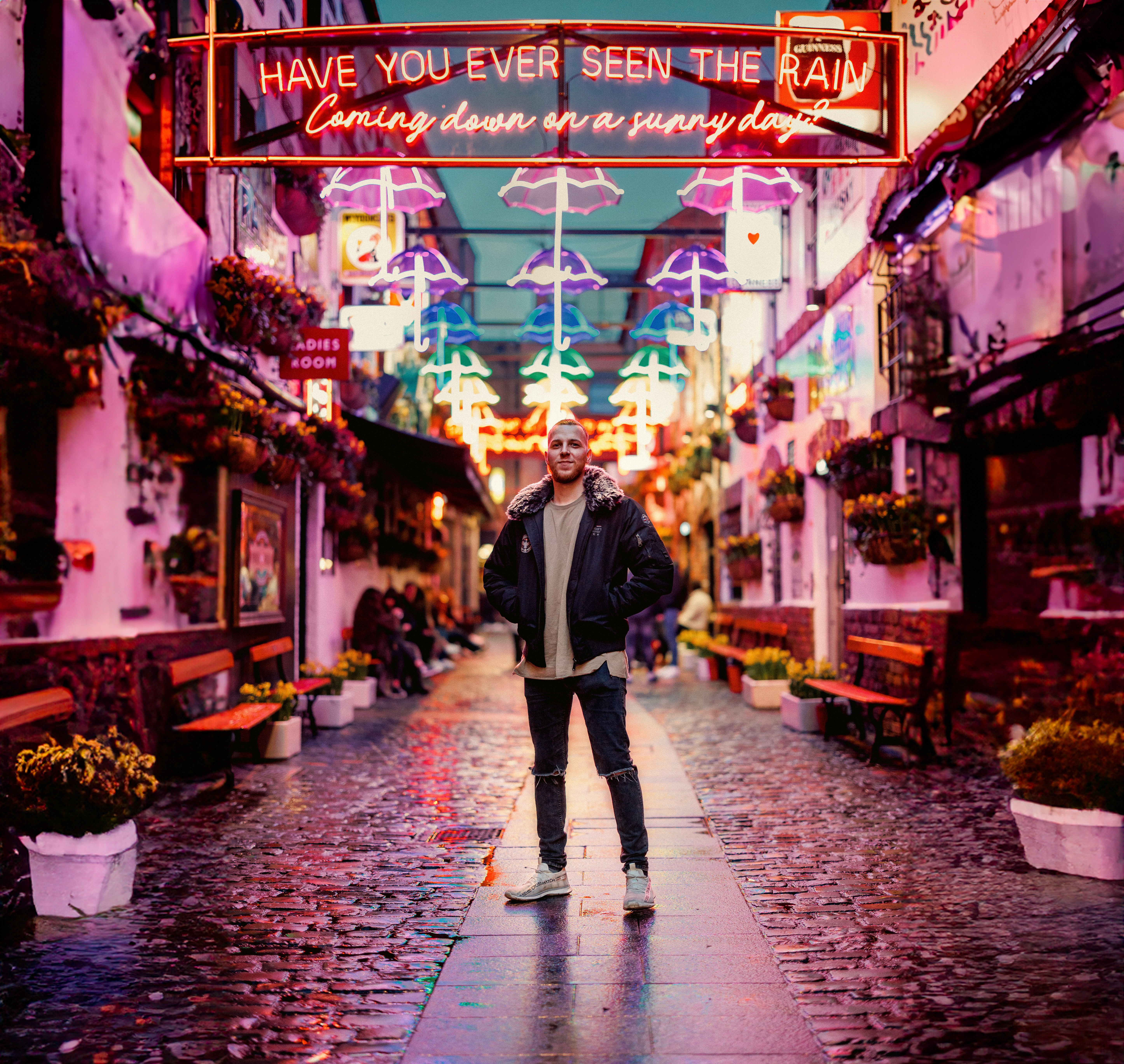 Egy színes, esernyőkkel díszített utcai jelenet neonfényekkel, középen egy kabátos férfi áll macskaköves úton.