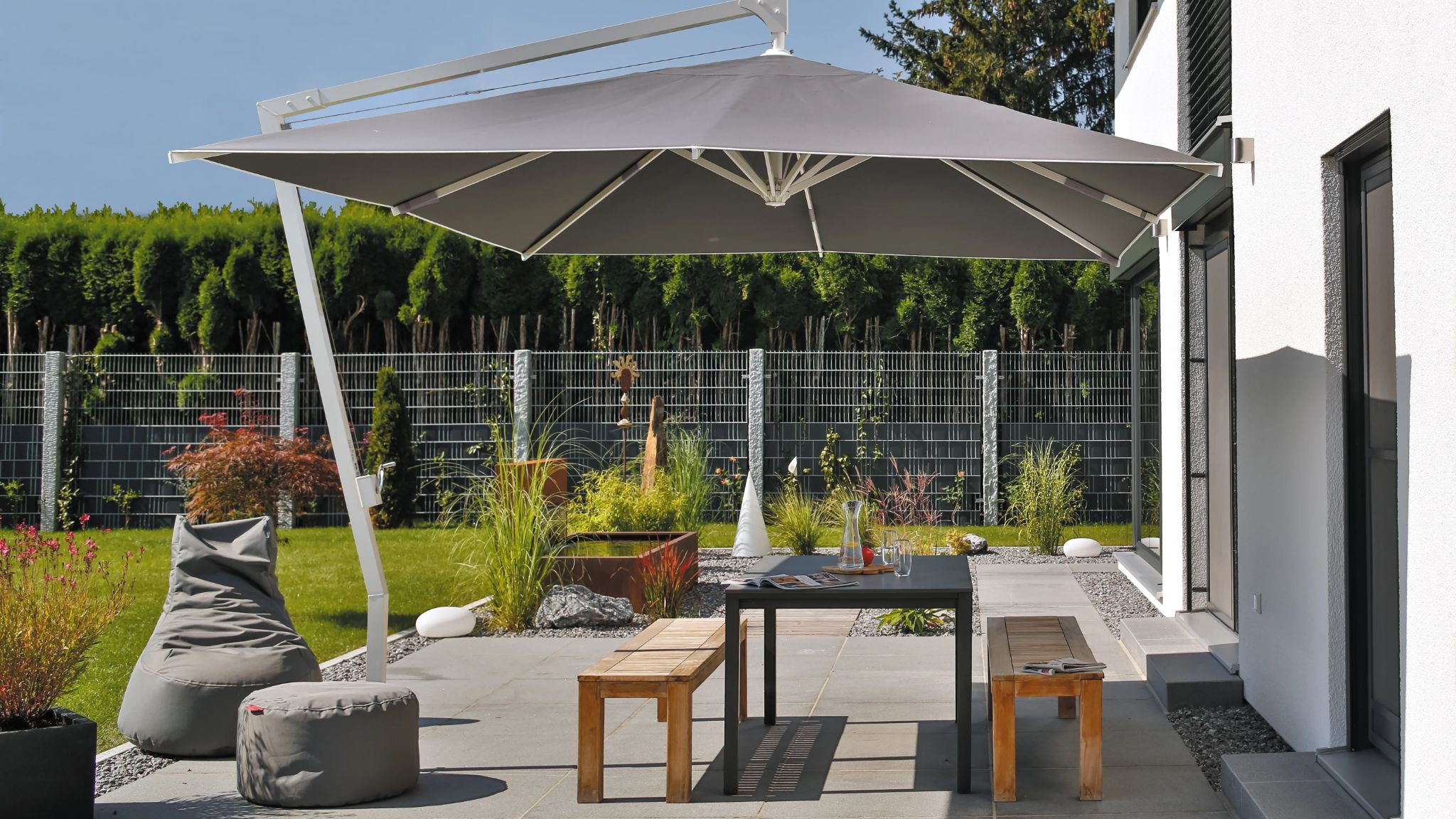 Großer, grauer Sonnenschirm mit Metallgestell über einer Gartenmöbelgarnitur aus Tisch, Bänken und Sitzsäcken auf gepflasterter Terrasse, im Hintergrund Gartengestaltung mit Zaun und Zierbäumen.