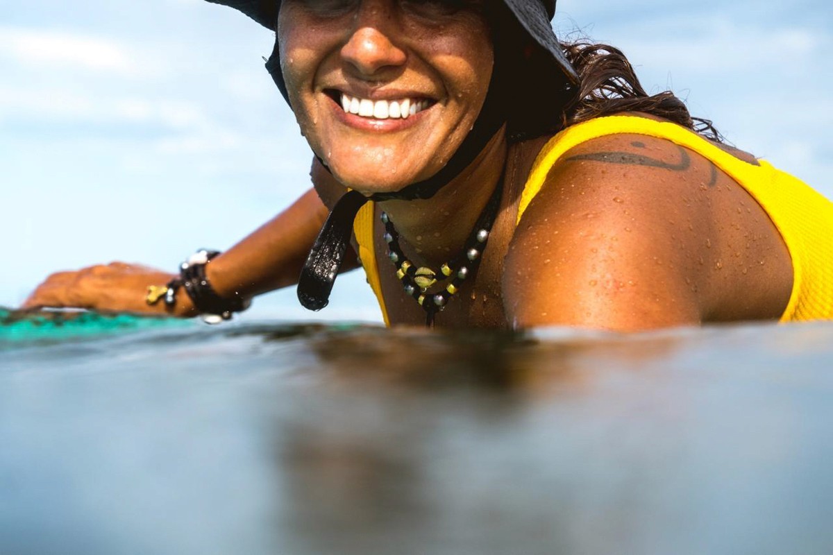 A person in a yellow swimsuit, smiling broadly while floating in the water, wears a wide-brimmed hat and beaded necklace, set against a clear blue sky background.