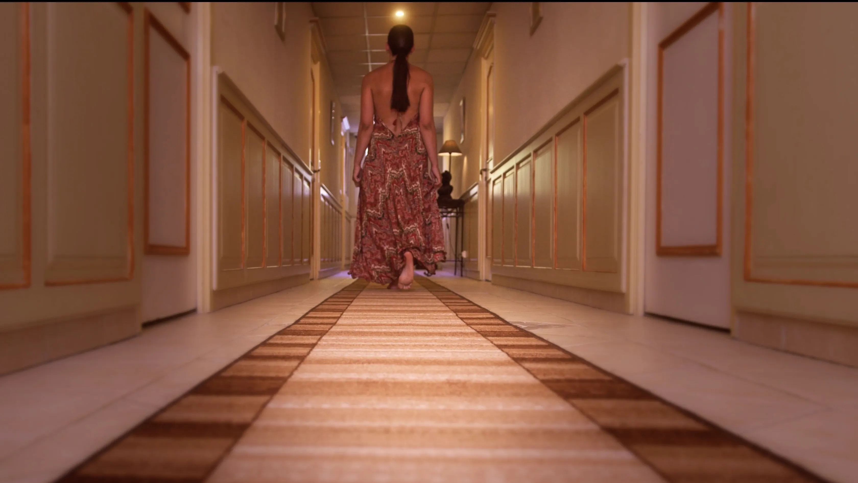 A person in a patterned dress walks down a softly lit, narrow hallway with beige walls and a striped carpet runner.