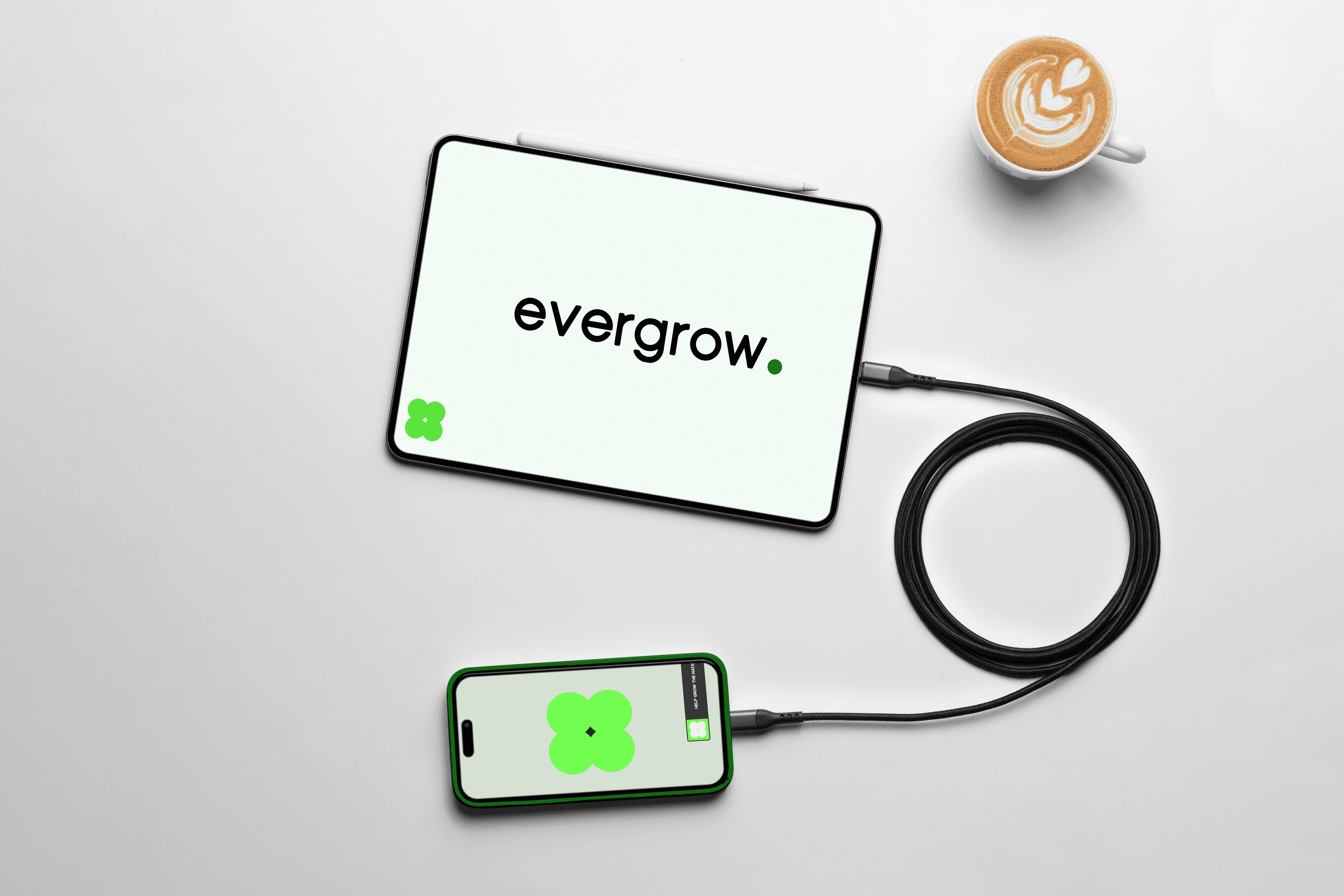 A tablet displaying the "evergrow" logo is connected to a smartphone with a matching green logo via a black cable, with a latte in a white cup placed nearby on a sleek, light-colored surface.
