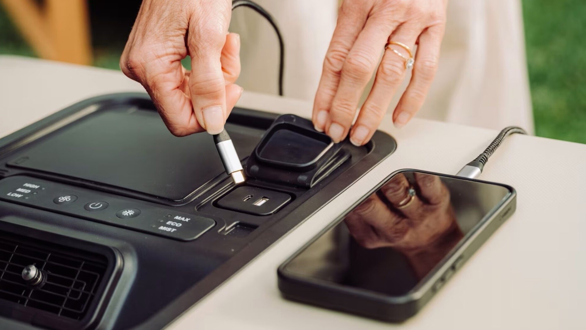 Hands plugging a USB-C cable into ports on a cooler's black control panel, with a phone charging beside it
