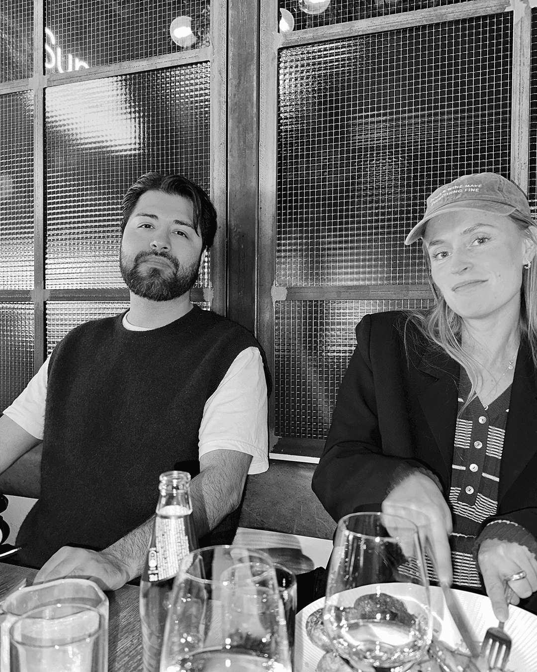 A black and white photo of a man with a beard and a woman wearing a baseball cap sitting together at a table.