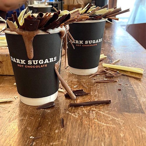 Dalawang tasa ng "Dark Sugars Hot Chocolate" na may umapaw na whipped cream at chocolate shavings sa isang kahoy na mesa.