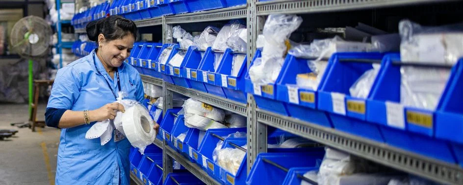 Mulher conferindo produtos em prateleiras com caixas organizadoras azuis em um estoque.