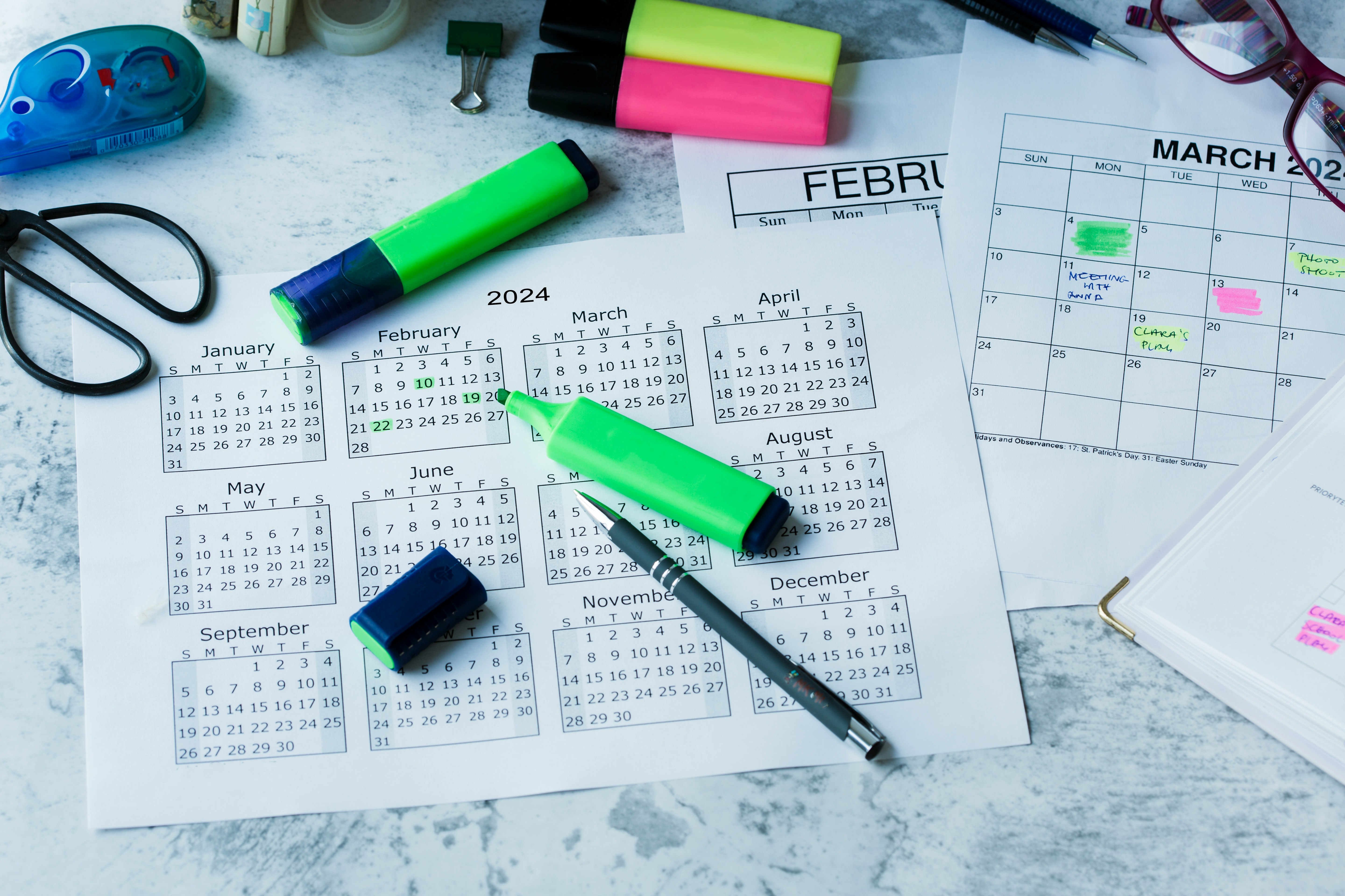 Year-at-a-glance calendar laid flat with green highlighters, pens, scissors, and a separate monthly planner page with handwritten meeting notes on a marble surface.