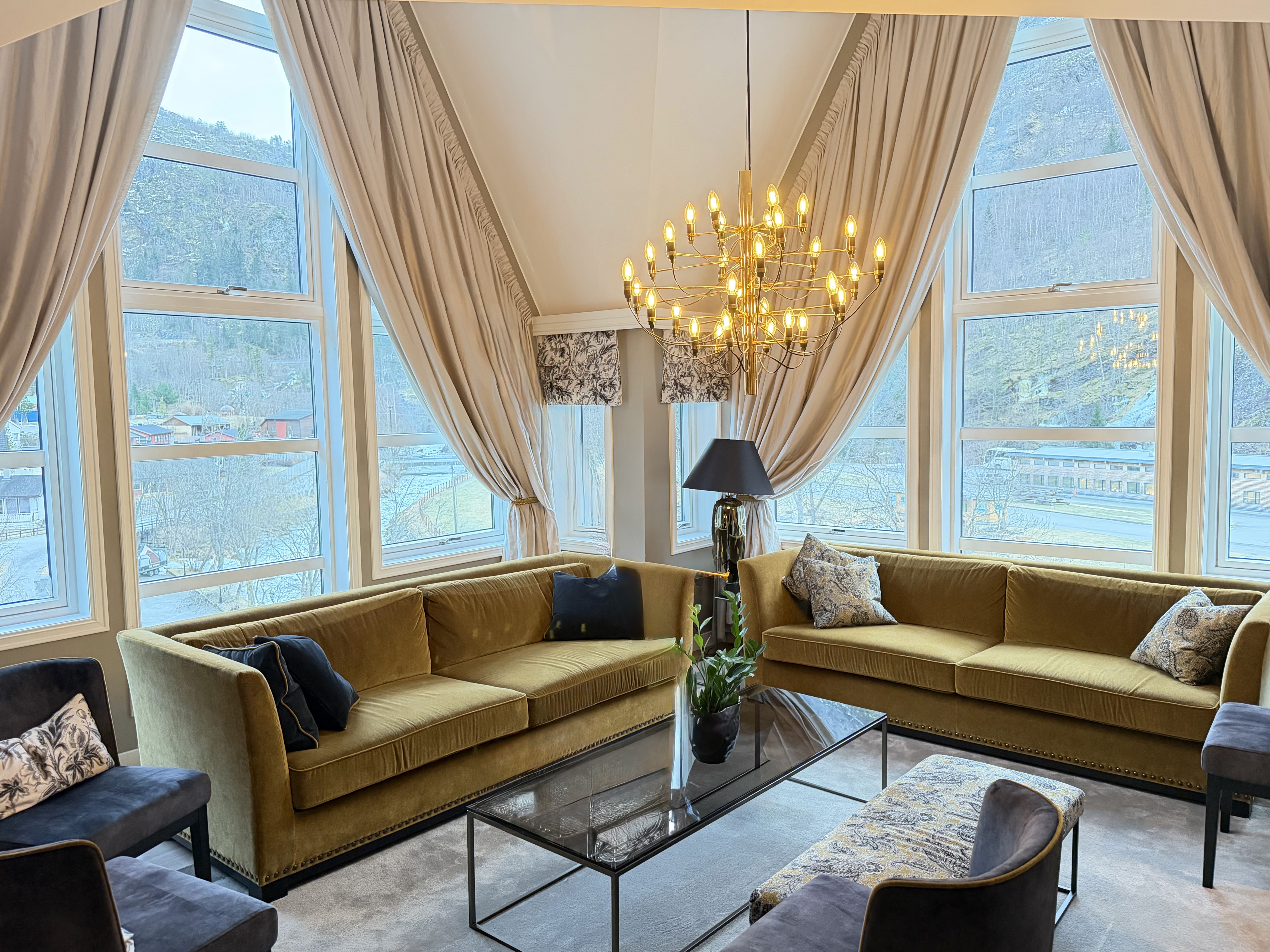 Luxurious bedroom with a stunning view features a plush bed adorned with patterned pillows, elegant floral curtains framing large windows overlooking green mountains and a serene lake, and a stylish pendant light hanging from the ceiling.