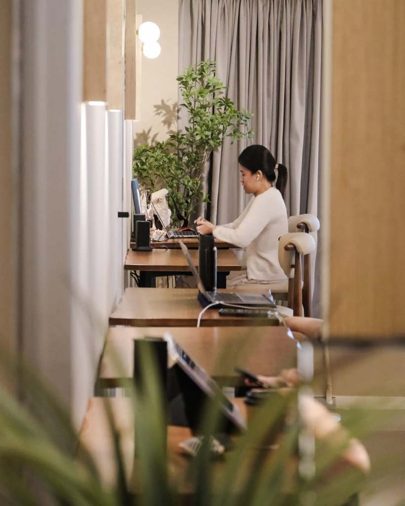 A woman working at a desk in a modern coworking space surrounded by greenery, with laptops and other office equipment visible.