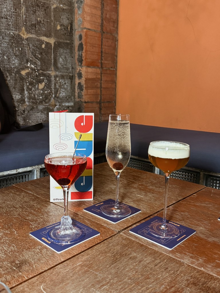 Three cocktails displayed on a wooden table in a bar, accompanied by a colorful menu. The drinks include a red cocktail garnished with a cherry, a clear sparkling beverage, and a golden drink topped with foam.