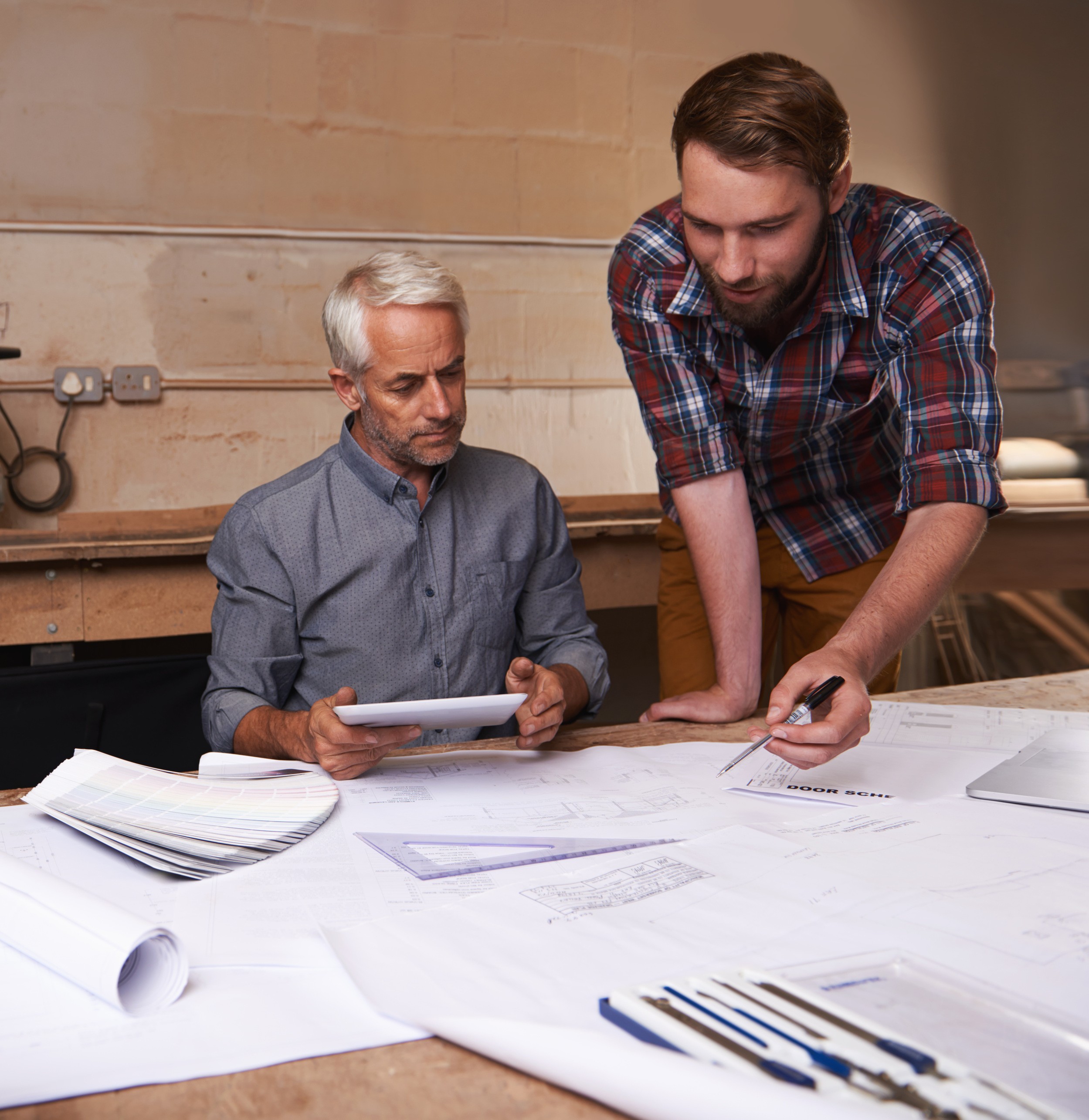 Two professional male designers collaborating over large architectural blueprints and a digital tablet in a studio.