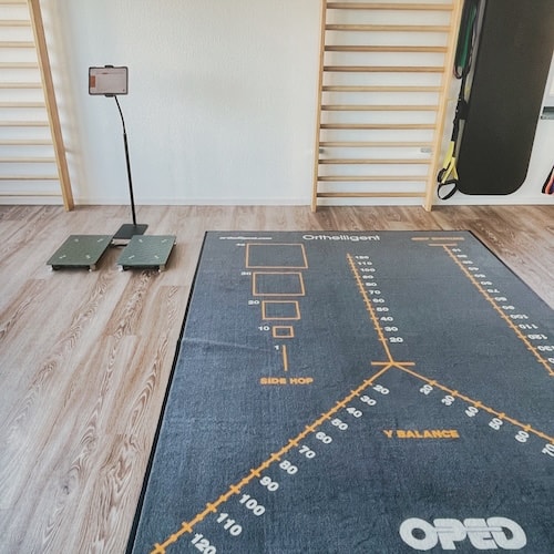 Zu sehen ist eine Y-Balance Testmatte mit aufgemalten Markierungen. Links daneben stehen die VALD Messplatten.