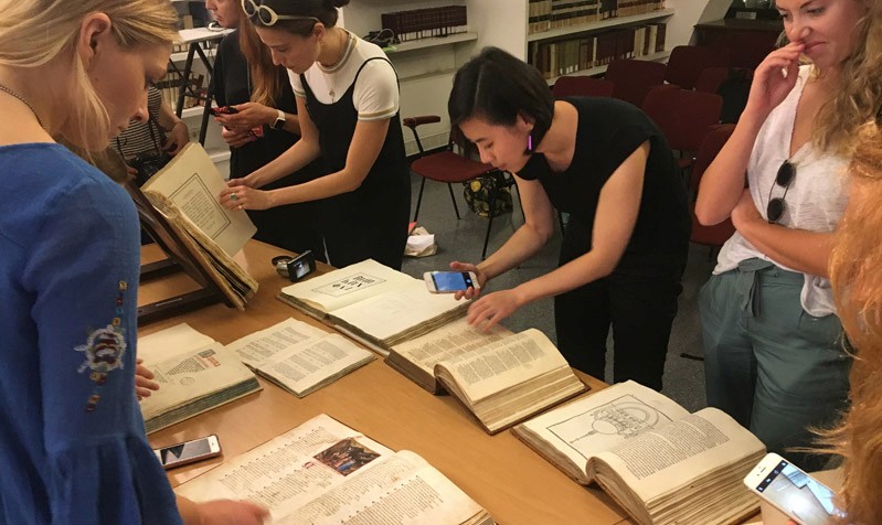 A photo of a woman taking pictures with her phone of some old book. There are also other people around the table checking some old books.