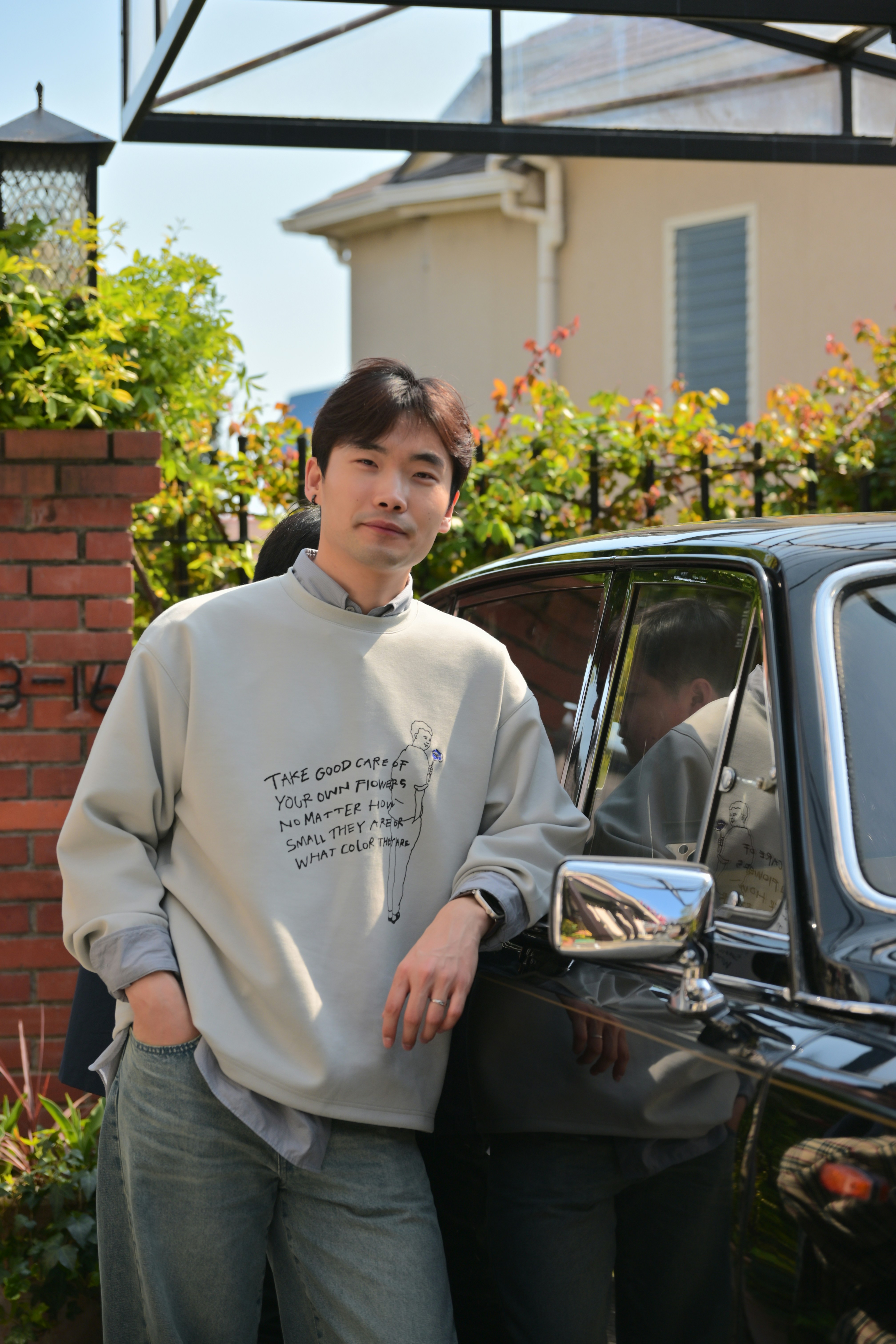 A person leans casually against a vintage black car parked next to a brick wall, wearing a light gray sweatshirt with text and light blue jeans, with a house and greenery in the background.