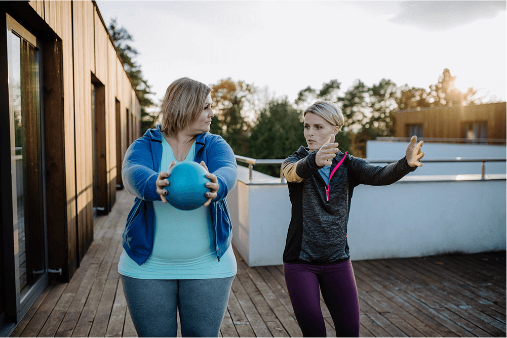 Two women exercising outdoors on a wooden terrace, with one holding a small blue medicine ball while the other gestures and gives instructions. Trees and building exteriors are visible in the background.