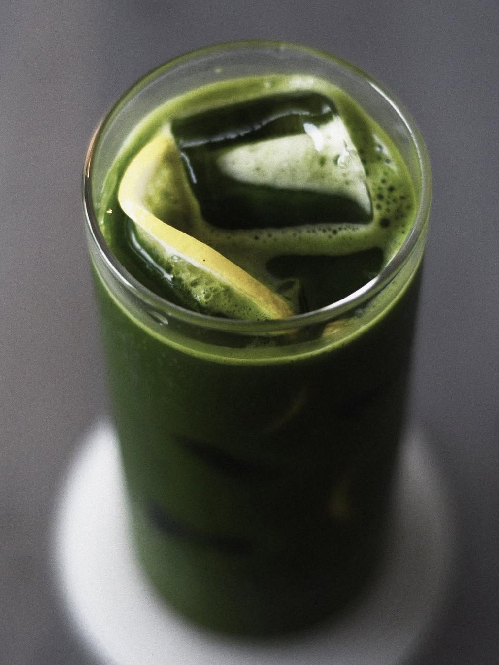 A tall glass filled with green beverage topped with ice cubes and a slice of lemon.