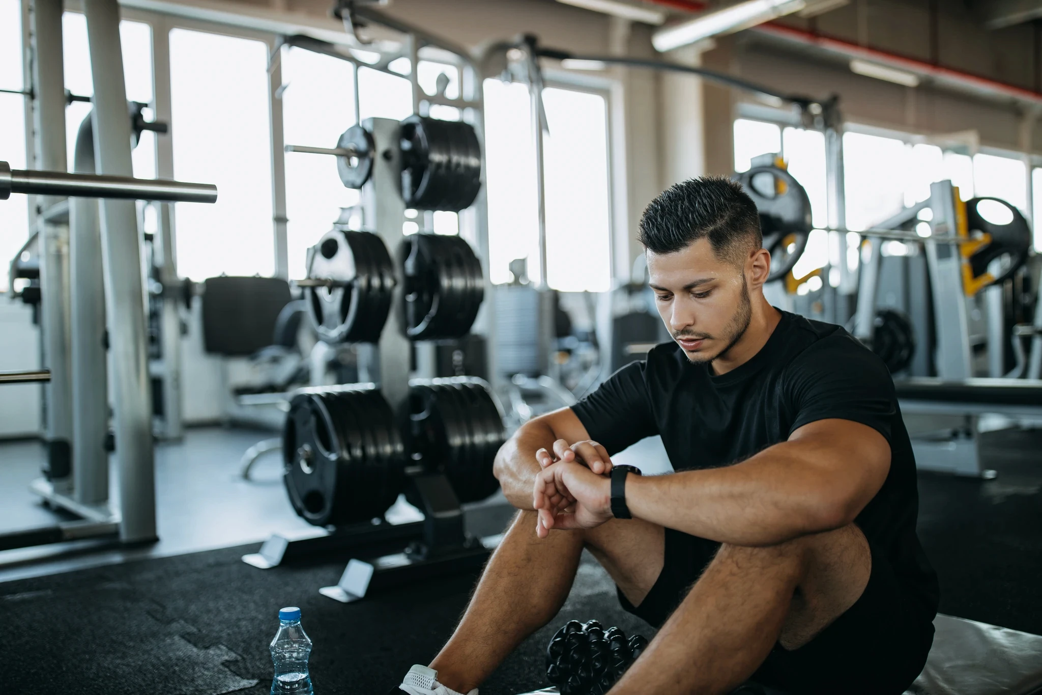 A man sitting on the gym floor, taking a rest between sets. He is looking at his smartwatch on his wrist, likely tracking his workout progress or timing his rest period.