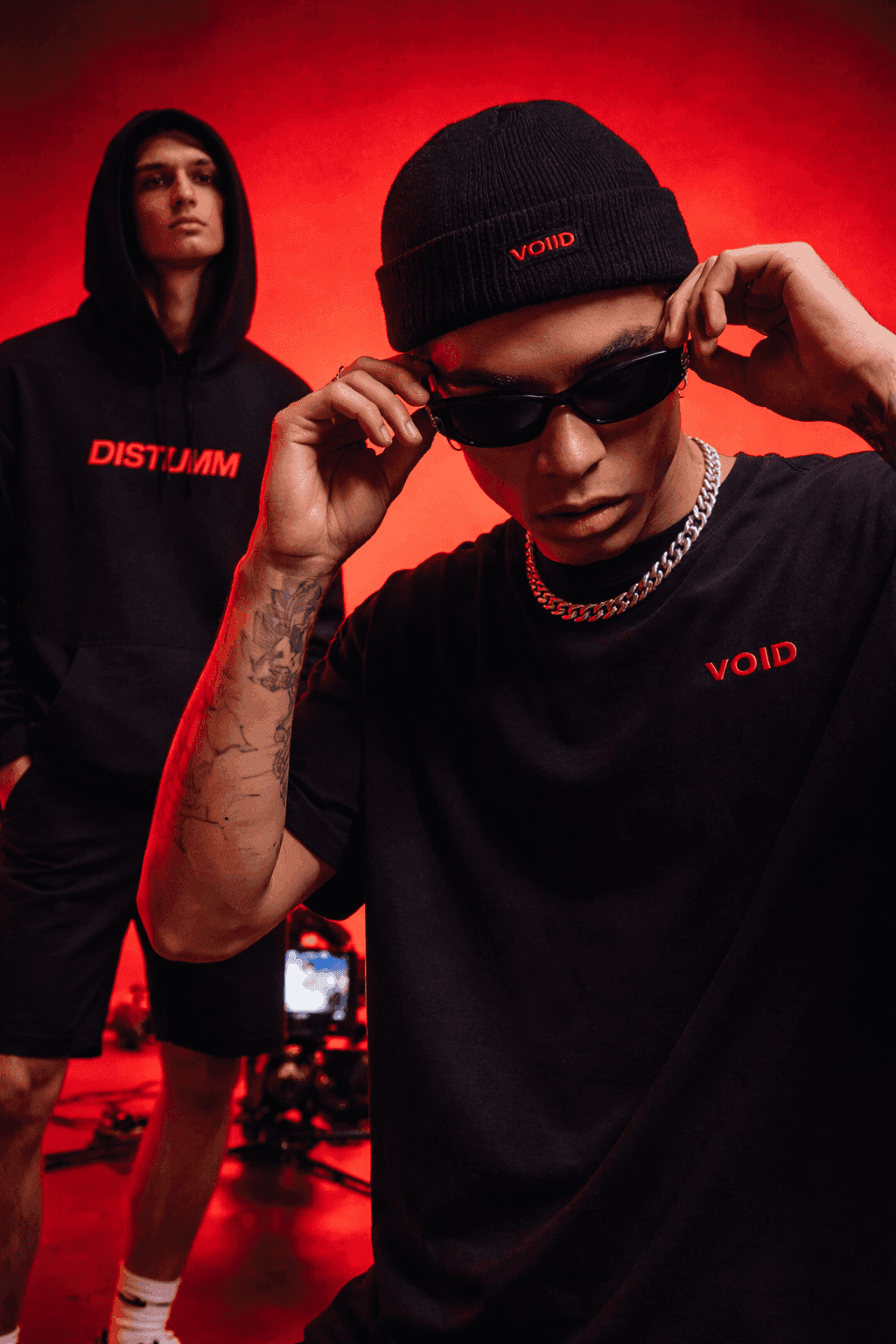 Two male models in black streetwear with red accents posing in studio with red lighting, one adjusting sunglasses in foreground