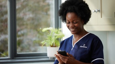 Healthcare worker in navy Florence scrubs smiling while using a smartphone near a window with natural light.
