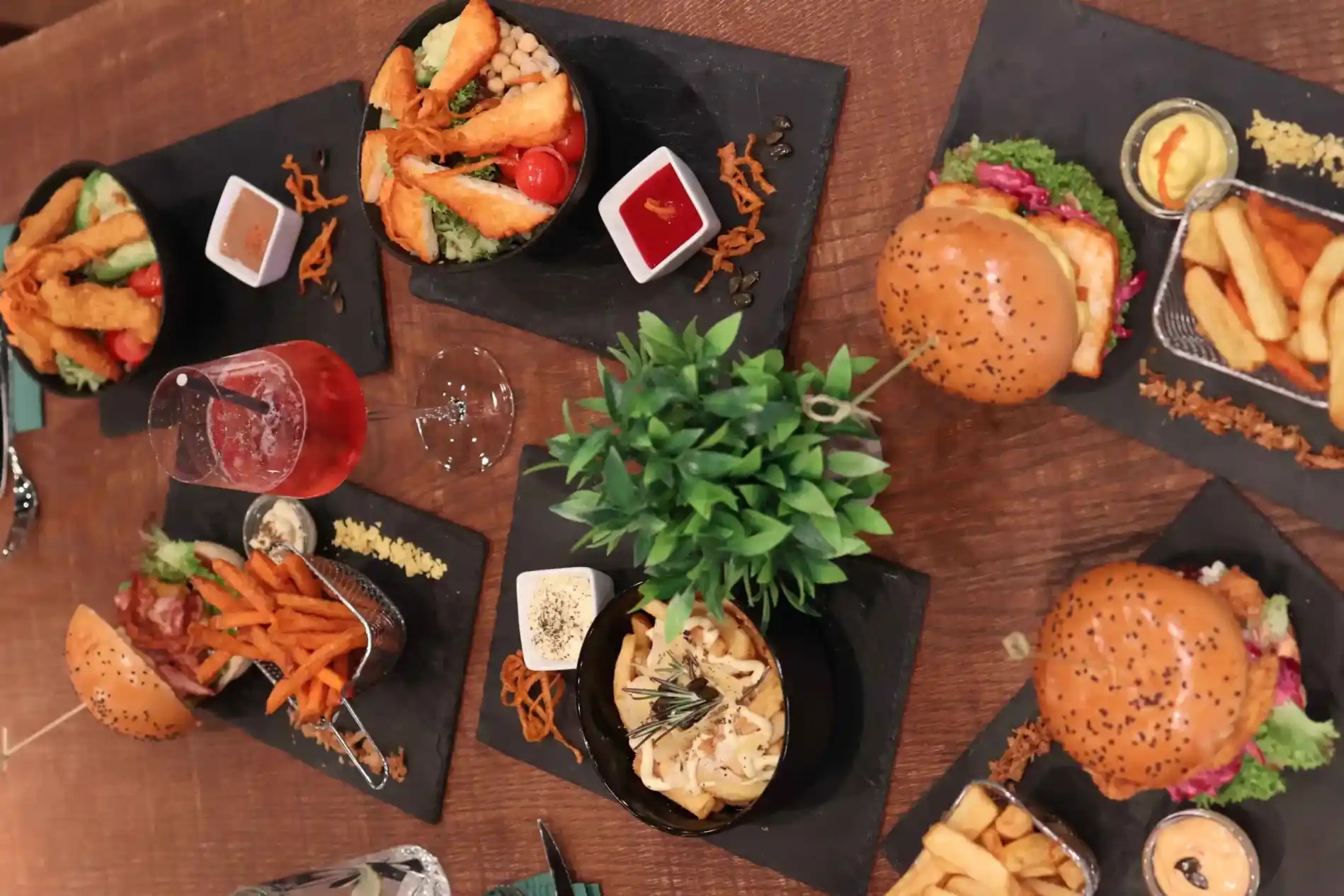 Overhead view of a kAlps Foodbar table spread with burgers, crispy fries, fondue dishes, a red cocktail, dipping sauces, and fresh garnishes arranged on slate serving boards.