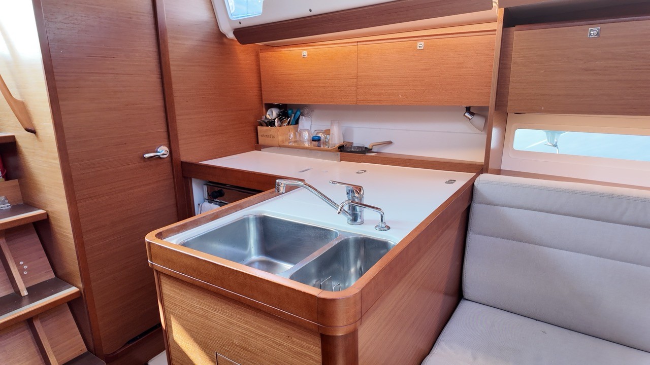 Interior view of the Dufour 350 GL's galley, showing a double stainless steel sink with a chrome faucet, teak cabinetry, an overhead storage locker, and a light gray settee to the right.